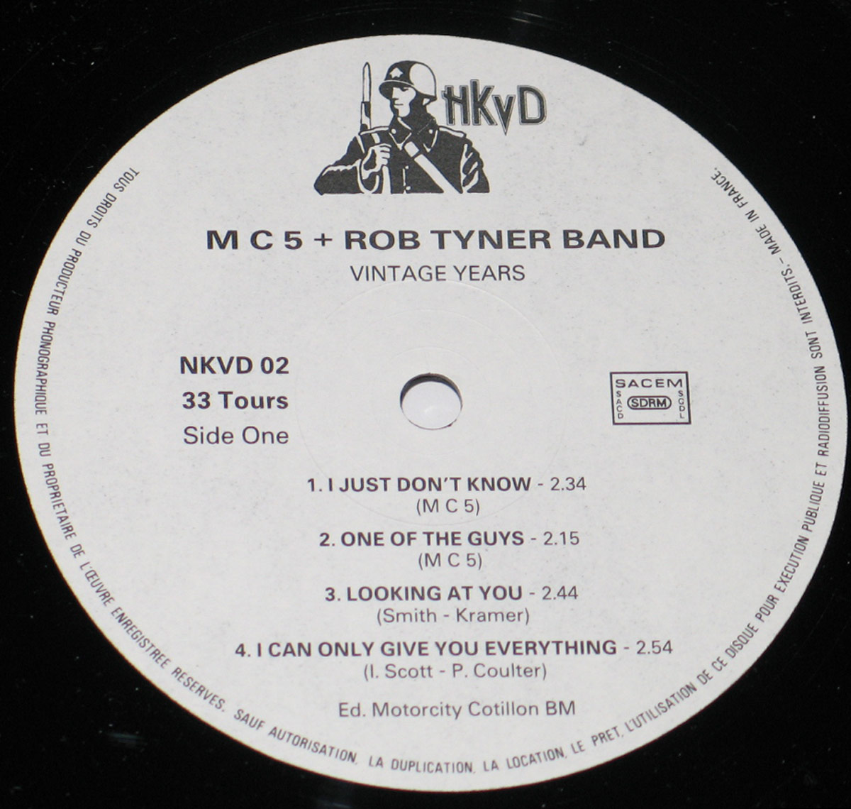 Close-up of the record label for Side One of 'MC5 + Rob Tyner Band - Vintage Years'. The label is white with black text, featuring the tracklist for Side One. At the top, there is an illustration of a soldier holding a rifle next to the 'hKyD' logo. The SACEM logo appears on the right side, indicating French pressing. The label also includes copyright and production information.