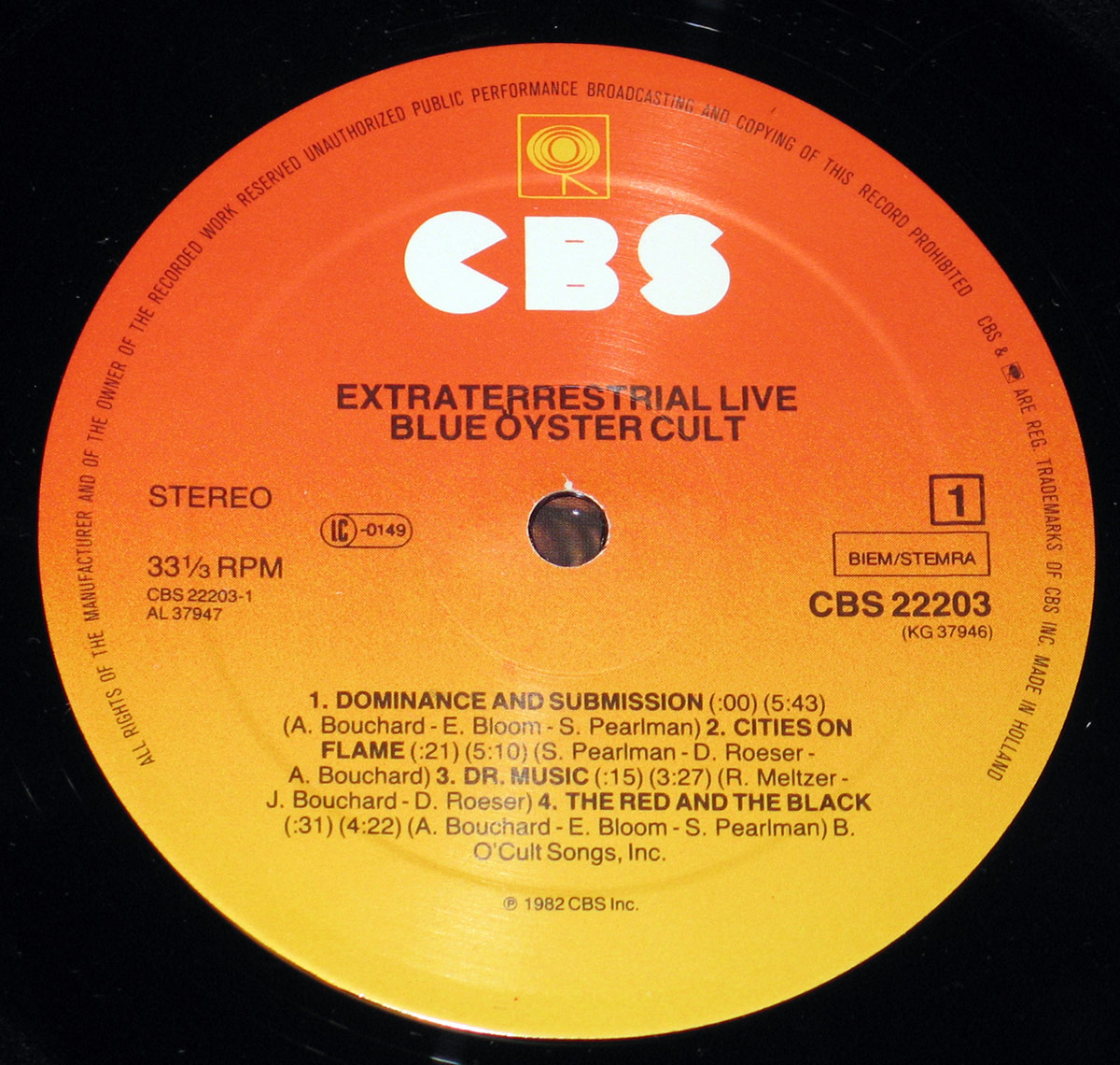 Close-up of the CBS record label on Blue Öyster Cult's 'Extraterrestrial Live' vinyl album. The label features a bold orange-to-yellow gradient background with the CBS logo at the top. Album title and artist name are printed in black uppercase letters. The tracklist for Side 1 is displayed, showing song titles, durations, and songwriting credits. Additional text includes catalog number CBS 22203, stereo format, and copyright details, with a central hole cut out for the spindle.