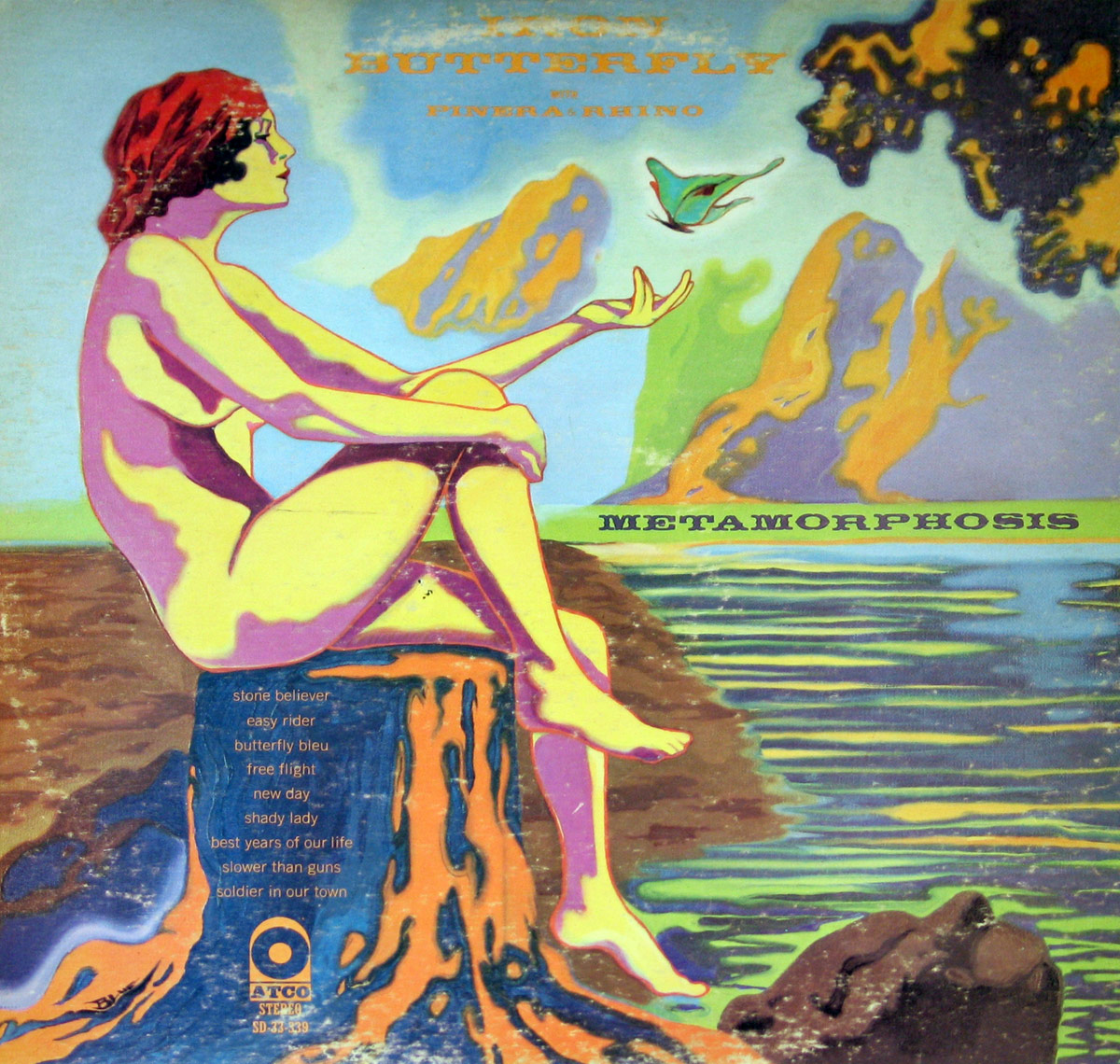 Back cover of Iron Butterfly's 'Metamorphosis' album. A psychedelic illustration featuring a nude, pink and yellow-hued woman with flowing red hair, seated on a melting tree stump. She extends her hand towards a fluttering butterfly. The surreal landscape includes stylized mountains, calm reflective water, and dripping organic forms in vivid blues, oranges, and greens. Album tracklist and Atco Records logo appear at the bottom.