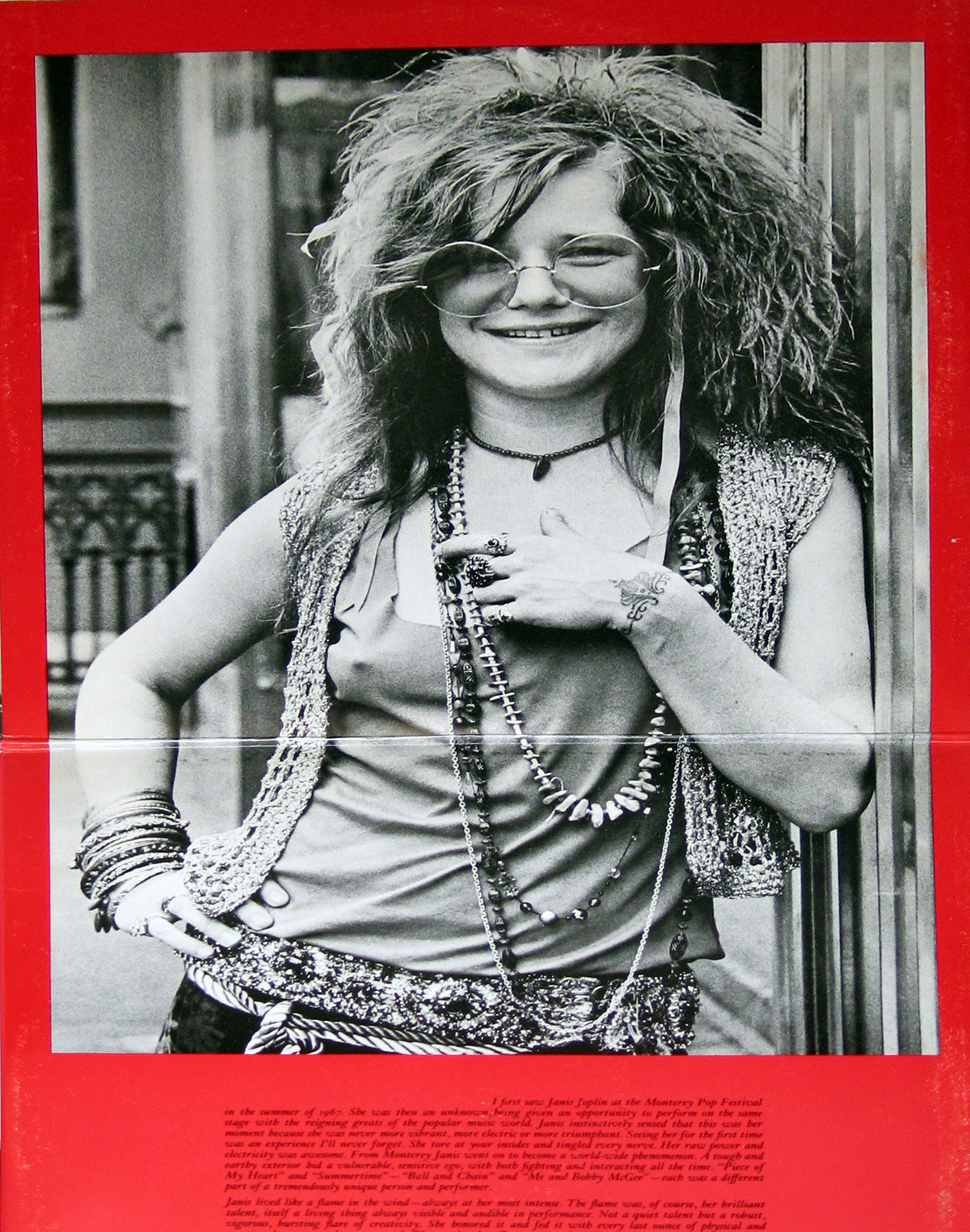 Inside gatefold poster of 'Joplin In Concert' featuring a radiant Janis Joplin leaning casually against a doorway, her wild hair framing her iconic round sunglasses. She wears layered necklaces, a crocheted vest, and a patterned sash, exuding bohemian energy. The lower red section contains a block of text reflecting on her impact and presence.