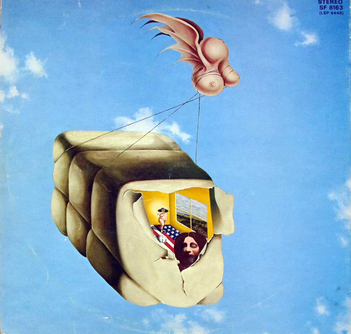 Surreal album cover art for 'Blows Against the Empire' by Paul Kantner and Jefferson Starship. The image shows a floating, cushion-like cube with an opening that reveals a room inside, where a person wrapped in an American flag and another figure near a window are visible. Above the cube, a pink, winged, breast-shaped object is tethered by strings, appearing to lift or guide the cube. The background is a blue sky with scattered clouds, creating a surreal, psychedelic scene.