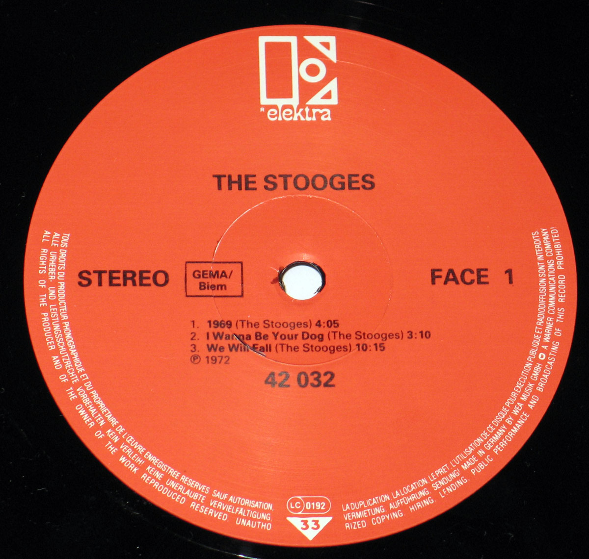 Close-up of Side One label from The Stooges' self-titled album, German Elektra pressing. The label is red with white text, featuring the Elektra Records logo at the top. Below, the album title 'The Stooges' is in bold capital letters, followed by track listings: '1969,' 'I Wanna Be Your Dog,' and 'We Will Fall.' Additional text includes 'STEREO,' 'GEMA/BIEM' rights information, and the catalog number 42 032. Small white text around the label's edge details copyright and legal notices.