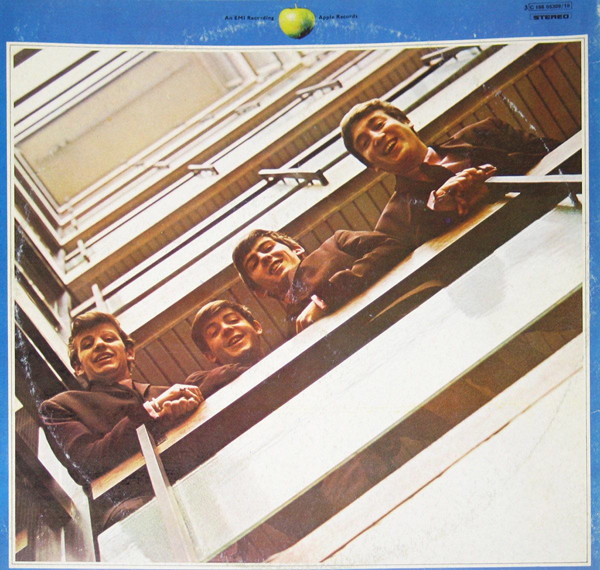 The back cover of The Beatles' '1967-1970' (Blue Album) features an earlier photograph of the band looking down from a stairwell. John Lennon, Paul McCartney, George Harrison, and Ringo Starr are dressed in matching dark suits, smiling with youthful energy. The metal railings and wooden beams form diagonal patterns, mirroring the front cover’s composition. The blue border remains, with an Apple Records logo at the top.