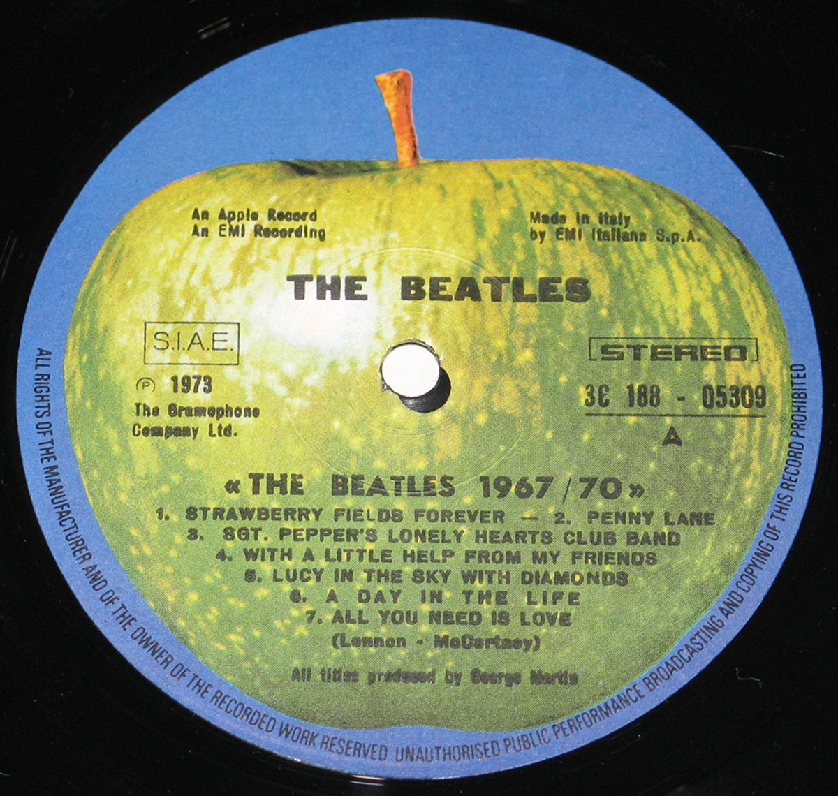 A close-up of the record label from The Beatles' '1967-1970' (Blue Album) Italian release. The label features the classic Apple Records design, with a green apple at the center against a blue background. The text displays the album title, 'The Beatles 1967/70,' along with track listings for Side A, including 'Strawberry Fields Forever' and 'A Day in the Life.' Additional information such as 'S.I.A.E.,' 'Stereo,' and 'Made in Italy by EMI Italiana' is printed around the label.