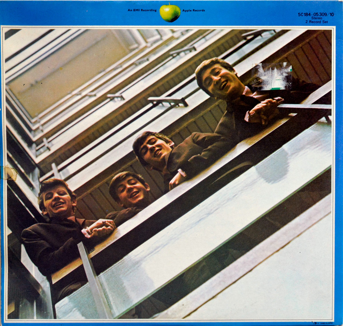 Back cover of The Beatles 1967-1970 Blue Album, featuring a youthful Beatles lineup leaning over a railing at EMI House in a recreation of their Please Please Me cover, framed by a blue border with Apple Records logo at the top.
