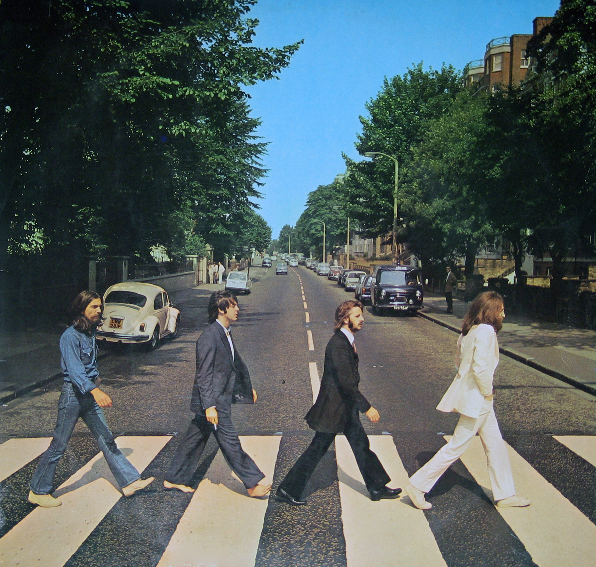 Album cover of The Beatles’ Abbey Road featuring the band members walking across a zebra crossing outside Abbey Road Studios in London. John Lennon leads in a white suit, followed by Ringo Starr in black, Paul McCartney barefoot with a cigarette, and George Harrison in denim. A white Volkswagen Beetle and other cars are visible in the background