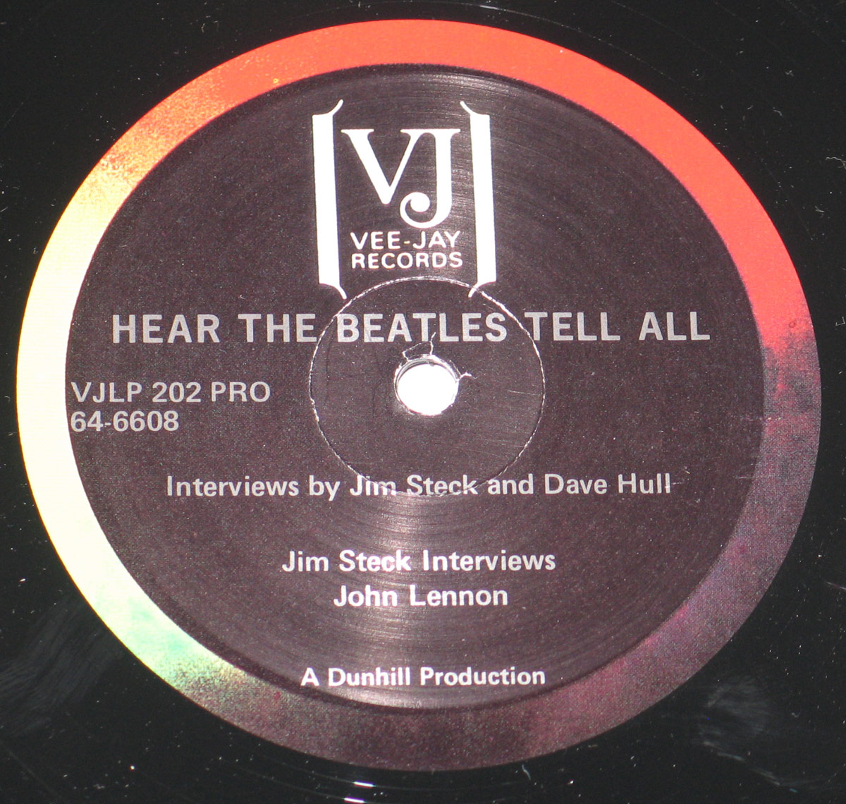 Close-up of the vinyl record label for 'Hear The Beatles Tell All' LP. Features the Vee-Jay Records logo at the top, album title in bold, catalog numbers, and interview credits. White text on a dark background with a red and yellow outer ring.