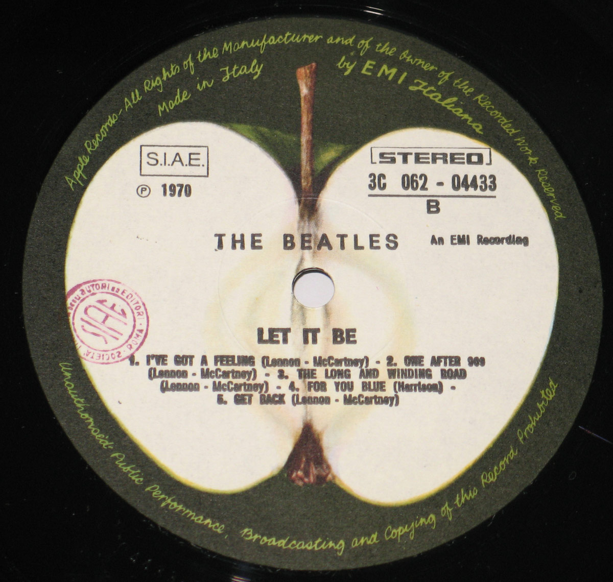 Close-up of the vinyl record label for The Beatles' 'Let It Be' Italian release. The design features the signature Apple Records label, depicting a halved green apple with a visible core. The album title and track listing for Side B are printed in bold black text, along with the catalog number '3C 062 - 04433'. The text ‘An EMI Recording’ and ‘Made in Italy by EMI Italiana’ are also present. A red 'S.I.A.E.' stamp is on the left side.