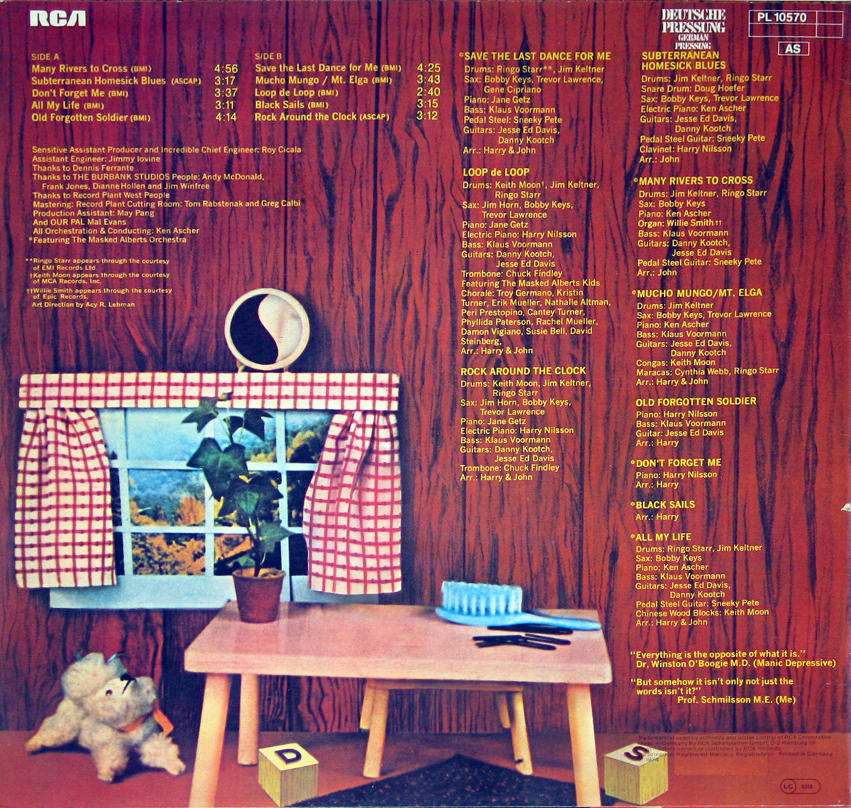 Back cover of 'Pussy Cats' by John Lennon and Harry Nilsson. The background resembles wood paneling, giving a cozy, rustic feel. A small window with red and white checkered curtains reveals a scenic outdoor view with greenery. A child-sized wooden table sits in the foreground, adorned with a blue hairbrush and a pair of black scissors. A white stuffed dog with a red ribbon collar sits on the floor, gazing up at the table. Wooden alphabet blocks with letters 'D' and 'S' are scattered on the carpet. Above the scene, yellow text lists the album’s track titles, credits, and production details. The RCA and Deutsche Pressing logos are visible in the upper right corner, along with catalog number 'PL 10570' and an 'A5' classification mark.