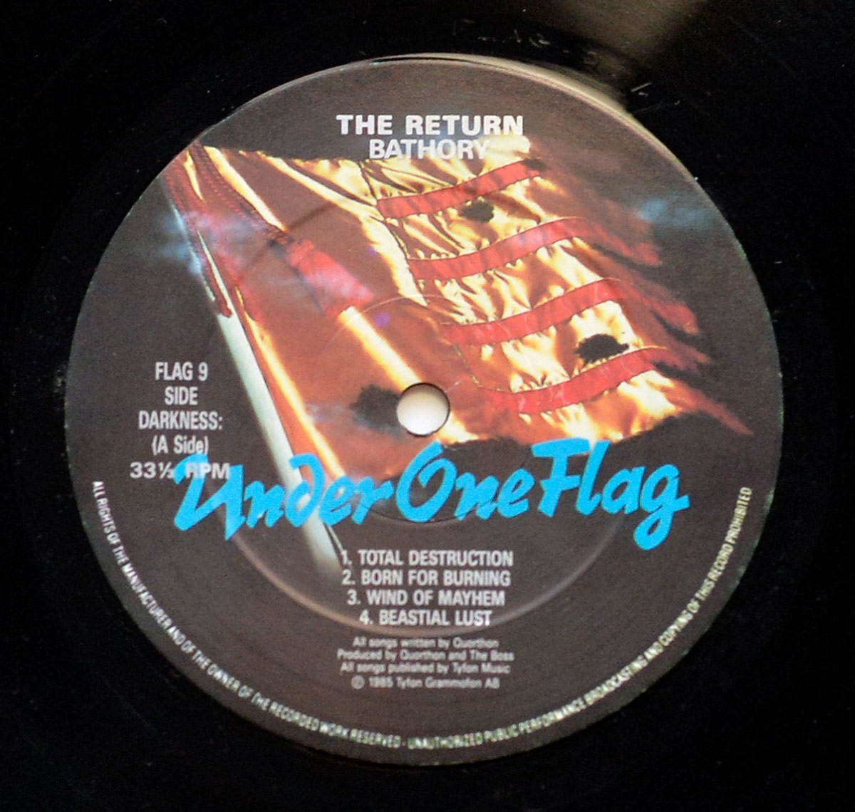 Close-up of the Side Darkness (A Side) record label of Bathory’s album The Return… of the Darkness and Evil. The circular label features the Under One Flag logo in bold blue script across the bottom. Behind it, a dramatic image of a red and gold flag waves with illuminated folds. At the top, the album title The Return and the band name Bathory are printed in white. To the left, catalog details read FLAG 9, Side Darkness (A Side), 33⅓ RPM. The track list is centered at the bottom half, showing Total Destruction, Born for Burning, Wind of Mayhem, and Bestial Lust. Production notes confirm all songs were written by Quorthon, produced by Quorthon and The Boss, published by Tyfon Music, © 1985 Tyfon Grammofon AB. The surrounding black vinyl grooves complete the presentation, framing the label’s stark design.
