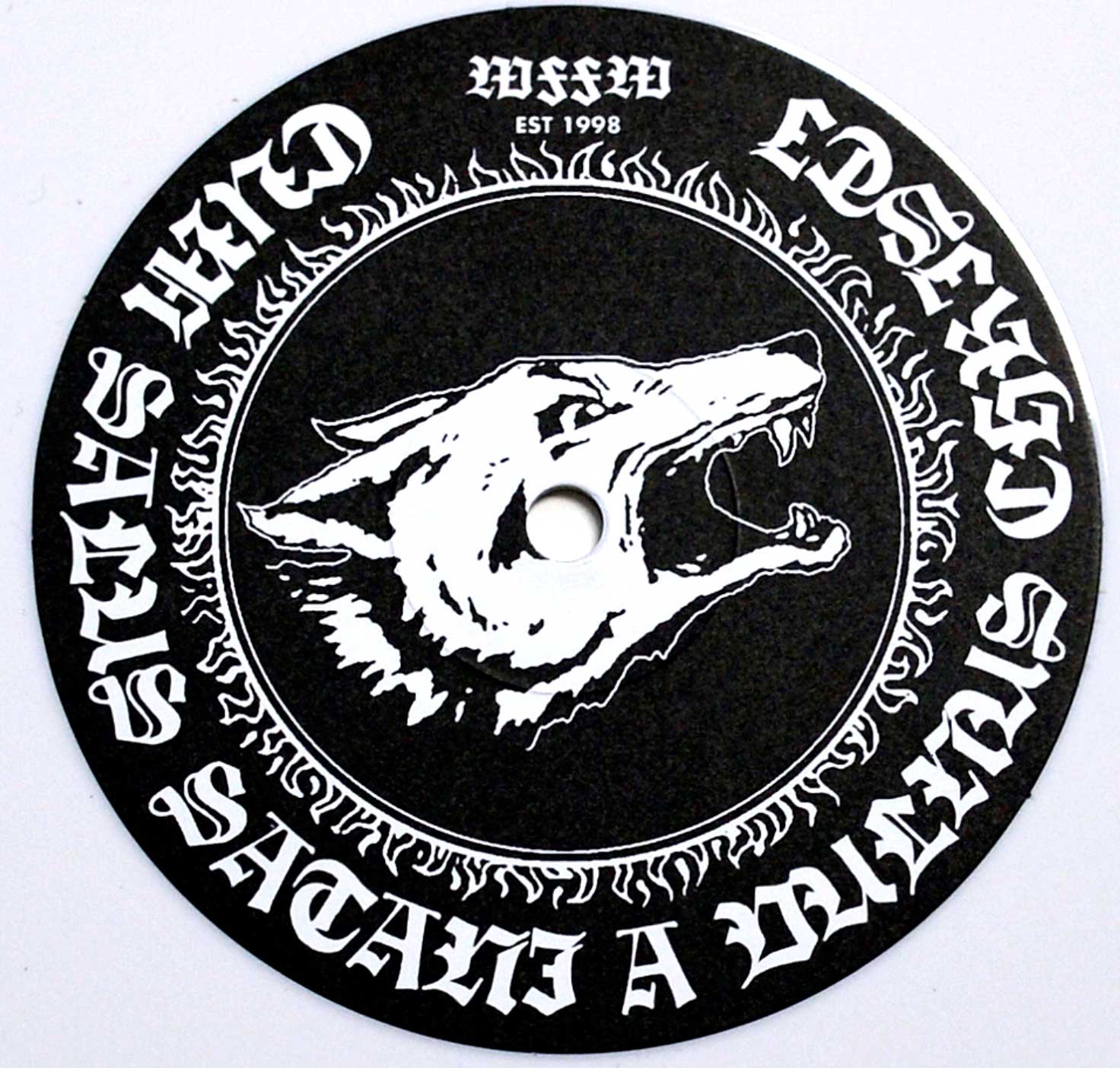 Close-up of the record label for Watain's 'Sworn to the Dark' White Vinyl edition. The design features a snarling wolf's head in a bold, high-contrast black and white style, encircled by a ring of flame-like details. Gothic-style text in an arc around the edge reads 'Satan's wolves and Lucifer's legions,' with the band's founding year, 'EST 1998,' and a runic inscription at the top.