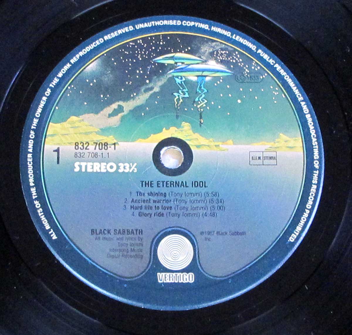 Close-up of Side One record label for Black Sabbath's 'The Eternal Idol' on Vertigo. It shows the tracklist with song durations, Vertigo swirl logo at bottom, catalog number 832 708-1, and a UFO-themed illustration above a stylized mountainous horizon.