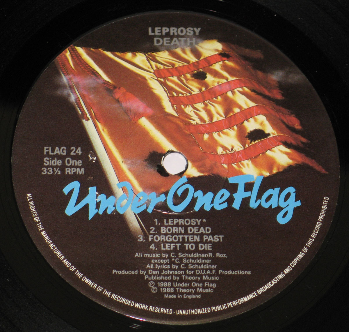 Record label of Death’s Leprosy album under the Under One Flag label. Features a stylized, torn golden American flag in the background. The album title Leprosy and band name Death appear in white at the top. The Under One Flag logo is centrally placed in bold blue, with additional details like catalog number, speed, and production credits along the edges