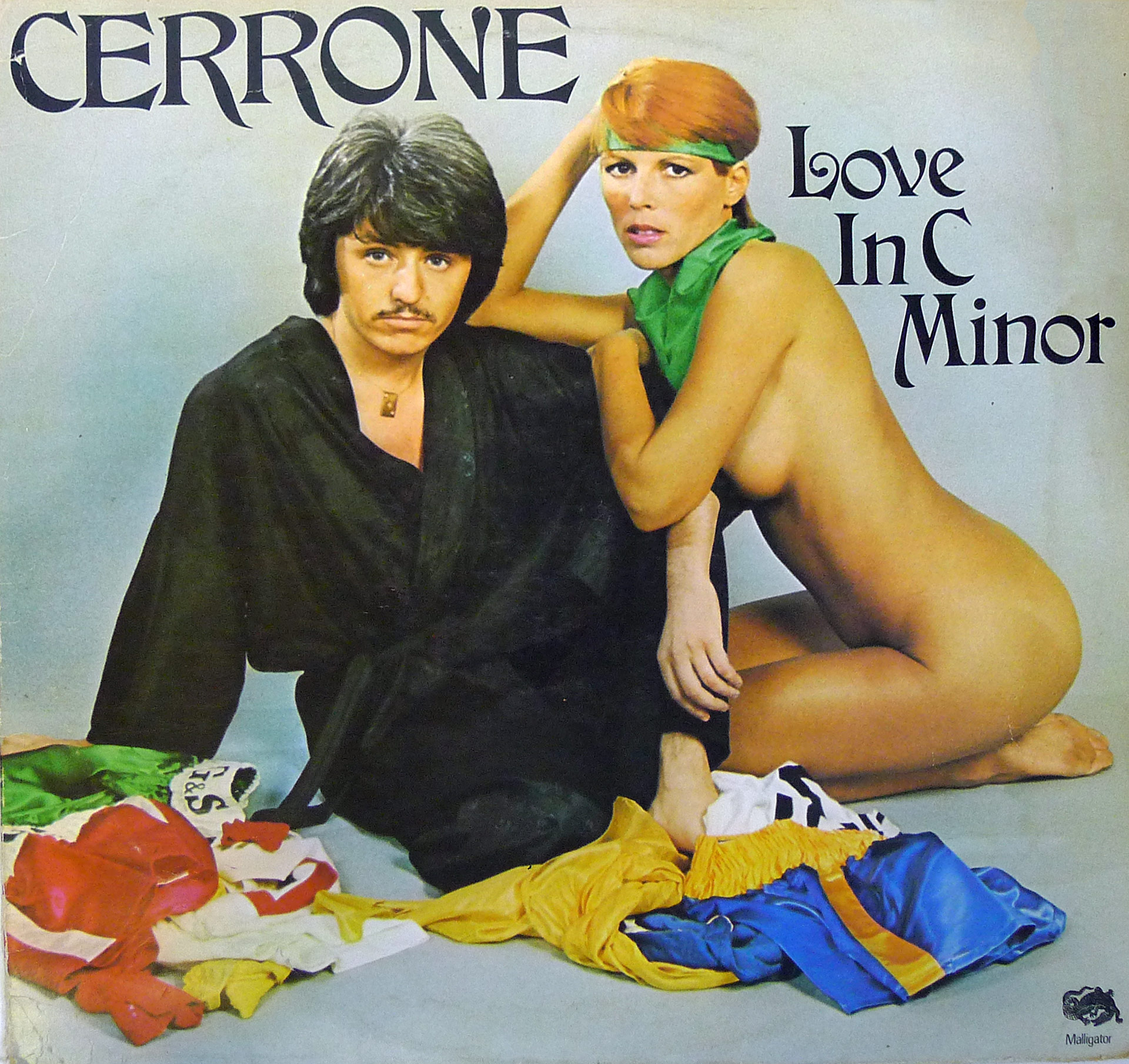 Album cover of Cerrone’s Love in C Minor featuring Marc Cerrone dressed in a black robe seated next to a nude woman with red hair and a green scarf, surrounded by scattered colorful fabrics, with bold stylized text displaying the artist’s name and album title.
