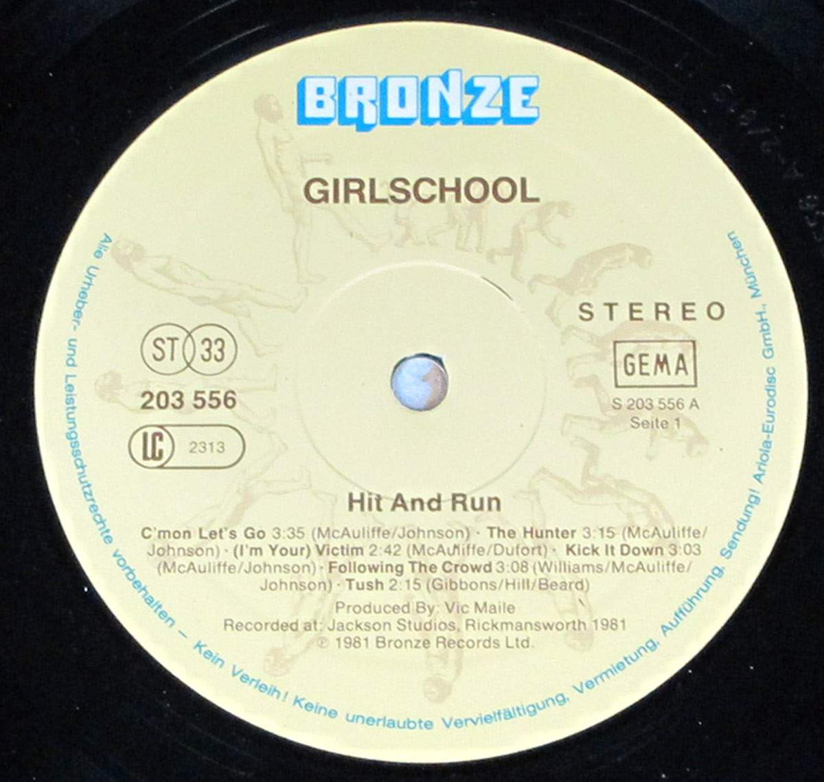 Close-up of the Side One record label for Girlschool - Hit and Run on a black vinyl LP. The cream label fills the centre, with large blue BRONZE logo at the top, GIRLSCHOOL beneath, catalog number 203 556, LC 2313, ST 33, GEMA, STEREO, and side marking S 203 556 A Seite 1. Track titles, credits to Vic Maile, and copyright text appear around the spindle hole and lower half.