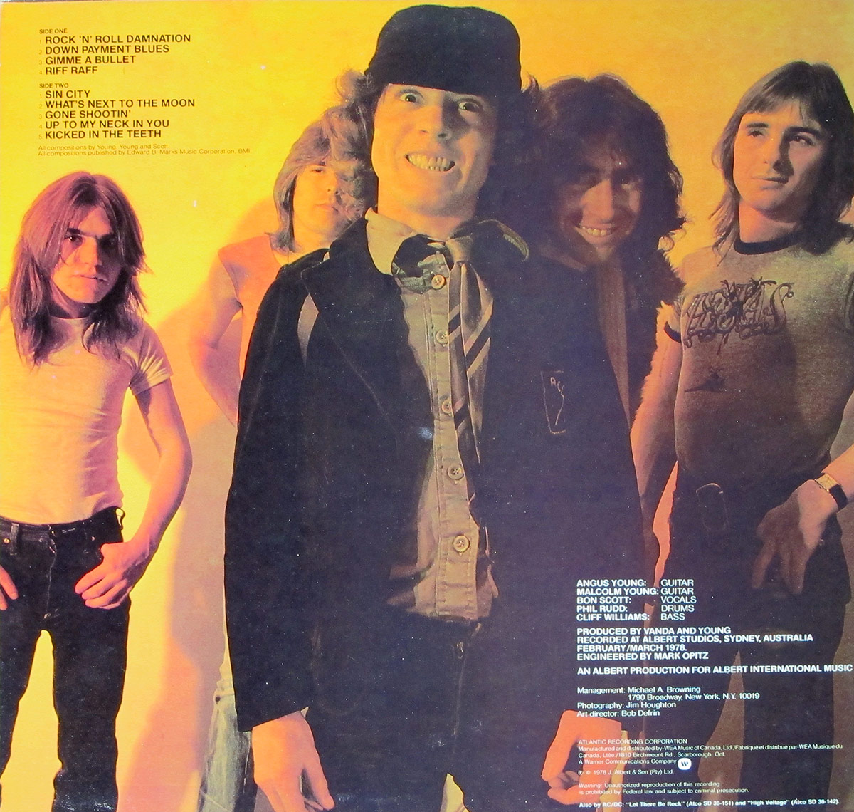 AC/DC Powerage album back cover featuring a group photo of the band members with Angus Young in his schoolboy outfit at the front, smiling energetically, and the rest of the band standing behind him against a vibrant yellow-orange background with production credits on the right