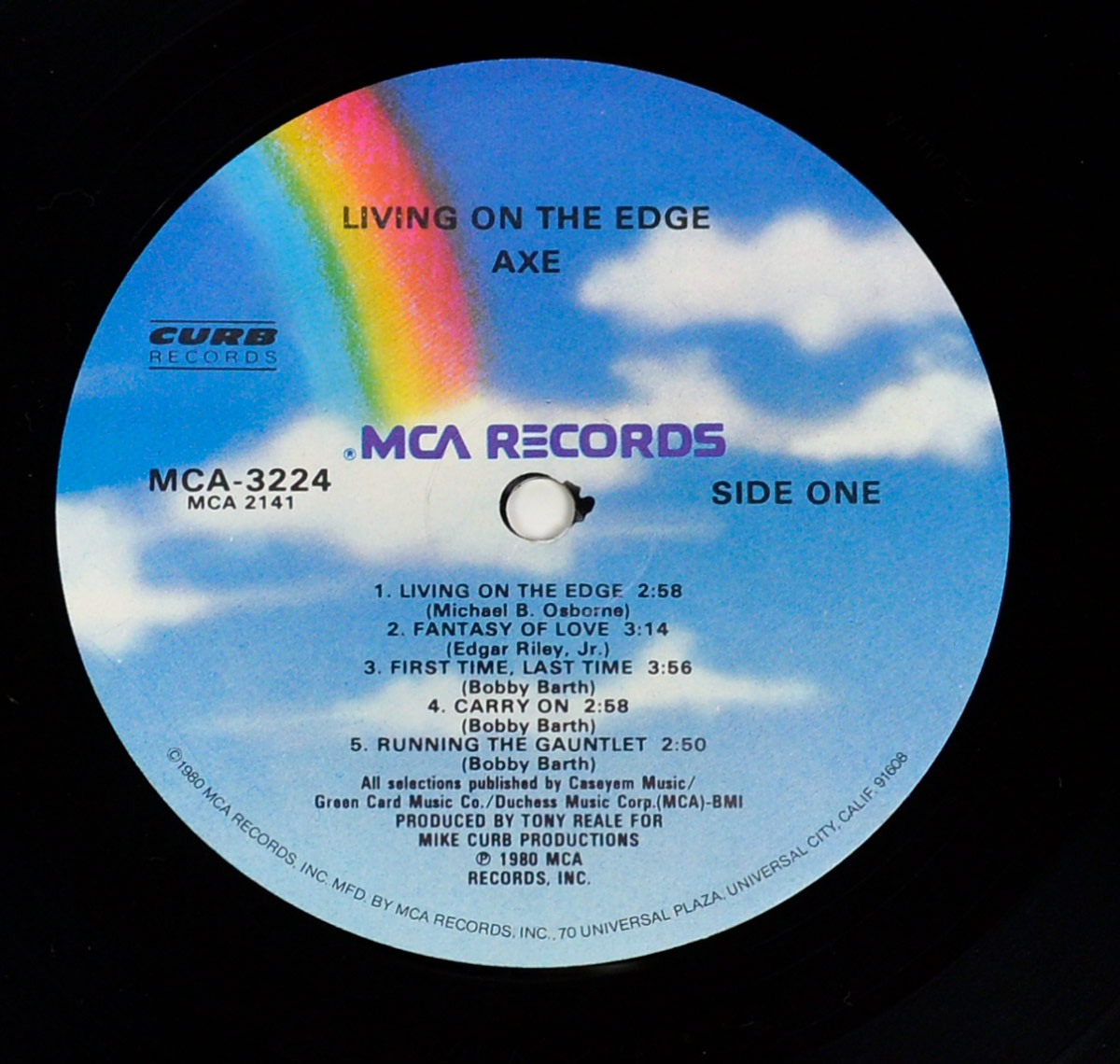 Side One record label of AXE’s 1980 album Living on the Edge, released on MCA Records. The circular label design features the signature MCA sky motif with soft blue background and fluffy white clouds, crossed by a vivid rainbow running diagonally from upper left to center. Text is printed in black with 'MCA RECORDS' in purple across the middle. The catalog number MCA-3224 is displayed on the left, alongside the Curb Records logo. The track listing is centered: Living on the Edge (Michael B. Osborne), Fantasy of Love (Edgar Riley Jr.), First Time, Last Time (Bobby Barth), Carry On (Bobby Barth), and Running the Gauntlet (Bobby Barth). Production credits note Tony Reale for Mike Curb Productions, with publishing and copyright info at the bottom edge.