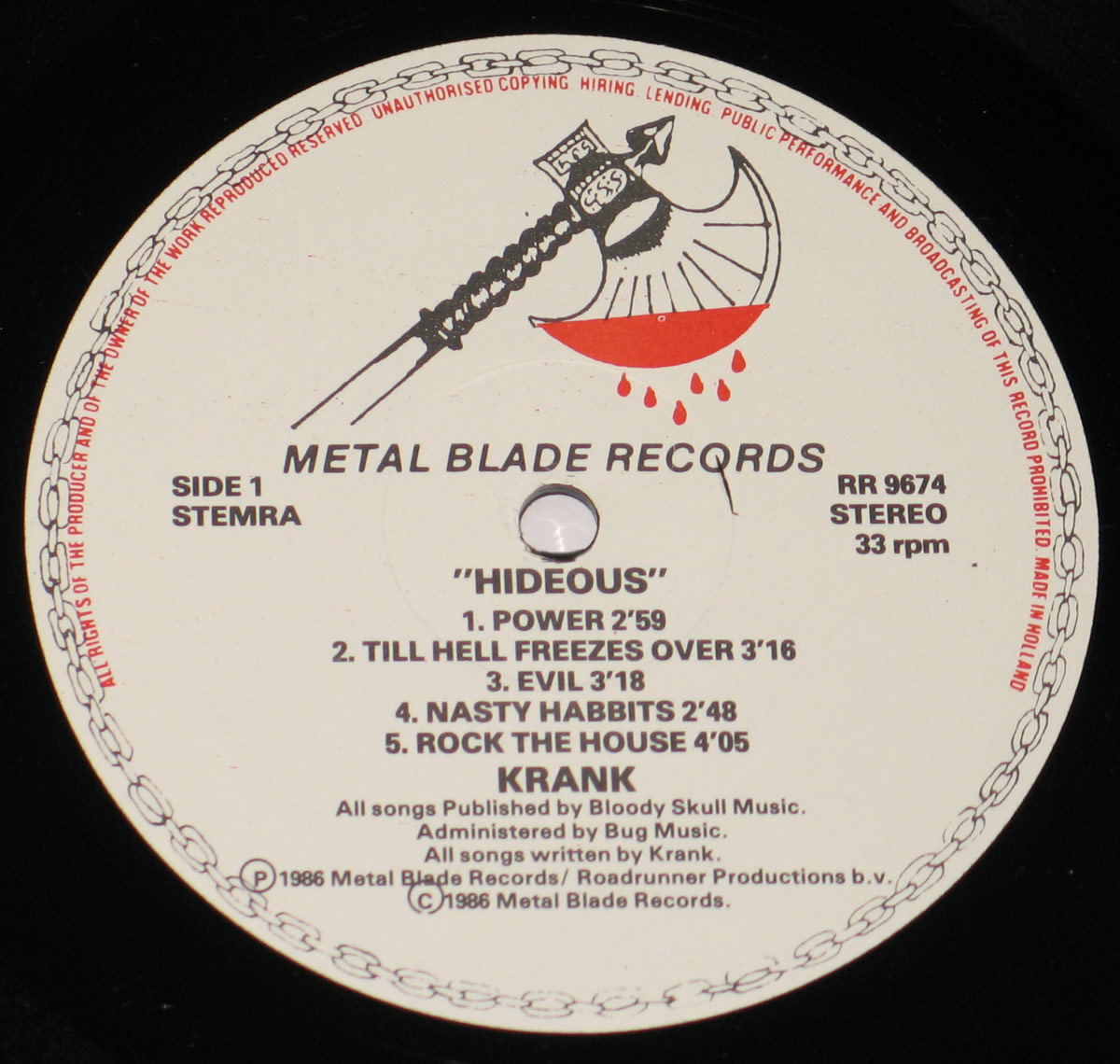 Close-up of the record label for Krank's 1986 album Hideous. The label is printed on a white background with black text and red accents. At the top, the Metal Blade Records logo is displayed, featuring a battle axe dripping with blood. Around the outer edge, a chain design encircles the label, with a red warning text stating unauthorized copying, hiring, lending, public performance, and broadcasting of the record is prohibited. The catalog number RR 9674 is listed to the right. The album title Hideous is printed in bold uppercase letters, followed by the track listing for Side 1, including song titles and durations. The band's name, Krank, is printed in bold, and a copyright notice at the bottom credits Metal Blade Records and Roadrunner Productions. The label also states that all songs were published by Bloody Skull Music and administered by Bug Music.
