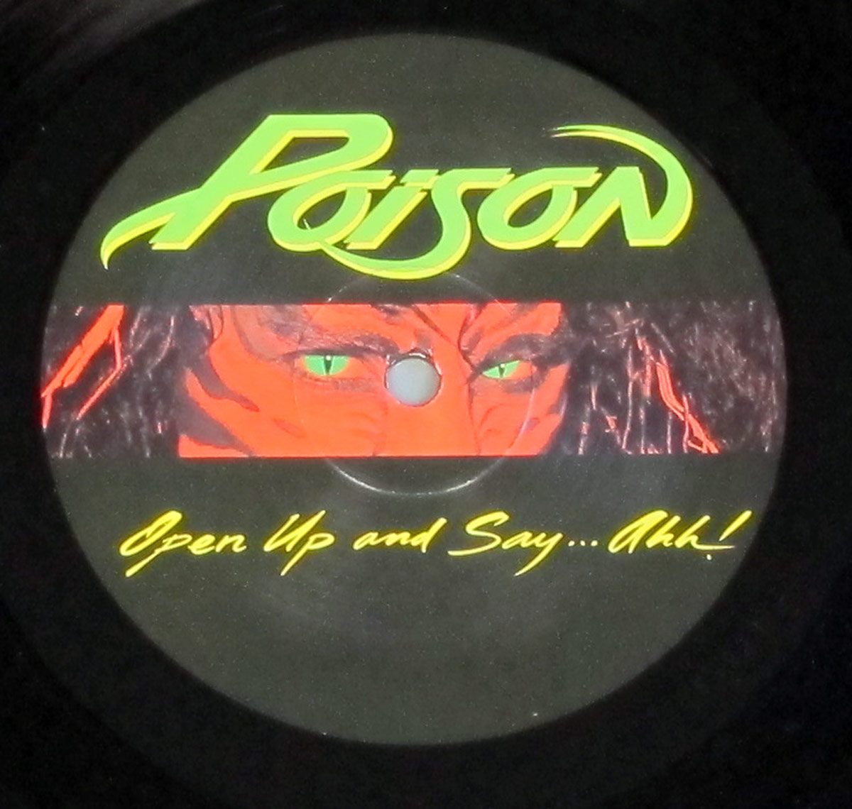 Record label of Poison’s 1988 album Open Up and Say… Ahh! featuring the band’s neon green logo at the top. Below the logo, a censored version of the album cover artwork is displayed, showing only the model’s green cat-like eyes framed by red and black tiger-striped face paint. The album title, ‘Open Up and Say… Ahh!’, is written in a yellow handwritten-style font at the bottom. The black background contrasts with the vibrant colors, maintaining the album’s bold aesthetic while incorporating the toned-down censored artwork