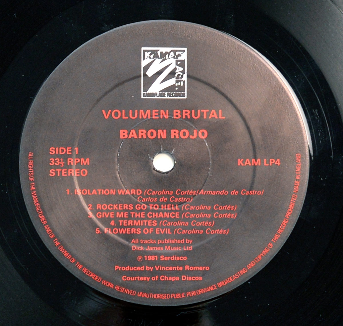 High-resolution close-up of Side One record label for Barón Rojo’s 1982 album Volumen Brutal, released on Kamaflage Records. The circular label is matte black with bright red text and the Kamaflage logo printed at the top. Around the spindle hole, in bold red letters, appear the album title Volumen Brutal and band name Baron Rojo. On the left margin are the words SIDE 1, 33⅓ RPM, and STEREO, clearly marking playback speed and format. The track listing for Side One follows: 1. Isolation Ward, 2. Rockers Go to Hell, 3. Give Me the Chance, 4. Termites, and 5. Flowers of Evil, with songwriting credits to Carolina Cortés and the de Castro brothers. Production credits note Vicente Romero as producer, courtesy of Chapa Discos, with publishing by Dick James Music Ltd. The catalog number KAM LP4 is printed on the right, and the rim text circles the label with legal copyright and reproduction warnings typical of early 1980s UK vinyl pressings.
