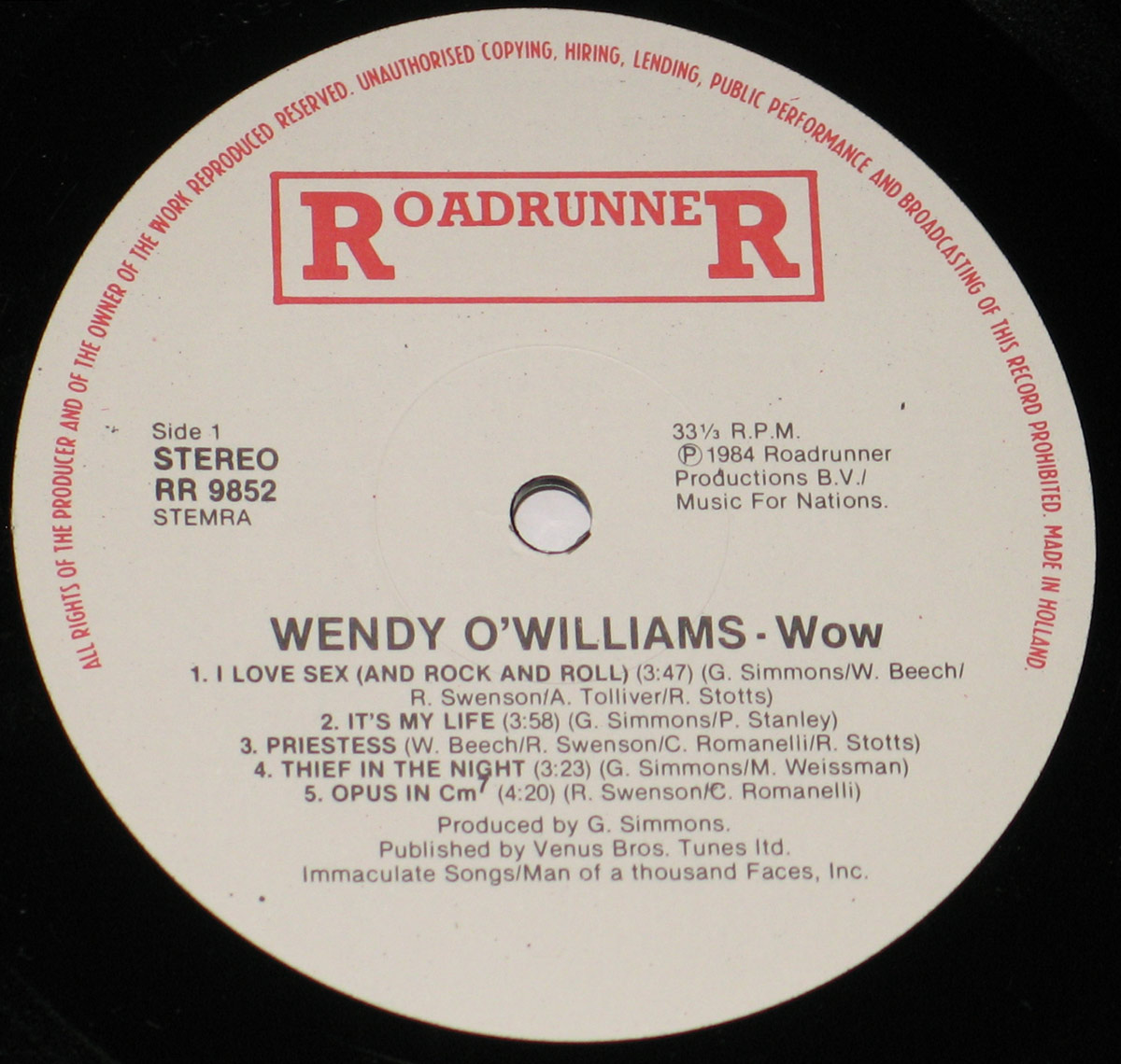 Close-up of the Roadrunner Records vinyl label for Wendy O. Williams – WOW. The label is white with red and black print, showing the Roadrunner logo at the top, catalog number RR 9852, Side 1 stereo designation, 33 1/3 RPM speed, and full track listing with songwriting credits. Fine surface marks and light reflections reveal this is an original pressed vinyl record, not a reprint.