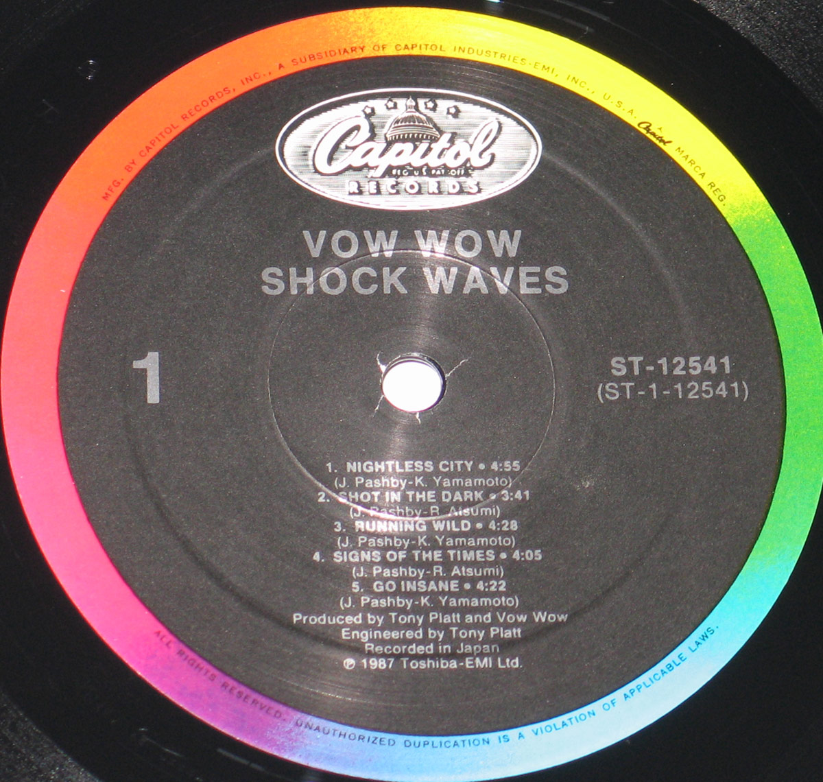 Close-up of the record label on Vow Wow's 'Shock Waves' vinyl LP, released by Capitol Records (ST-12541) in 1987 under Toshiba-EMI Ltd. 
        The black label features the Capitol Records logo at the top, with a multi-colored outer ring transitioning from red to yellow to green. 
        Track listings for Side 1, production credits, and copyright details are printed in white text.