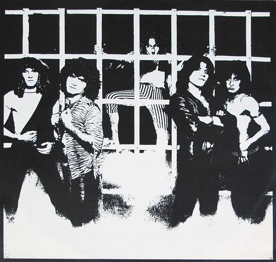 Black-and-white photo of the band Krokus posing in front of a barred, jail-like backdrop. Four band members stand in the foreground with intense expressions, while one member in striped pants is positioned behind the bars, adding depth. The high-contrast image has a gritty, rebellious vibe, highlighting the band’s hard rock attitude.’
