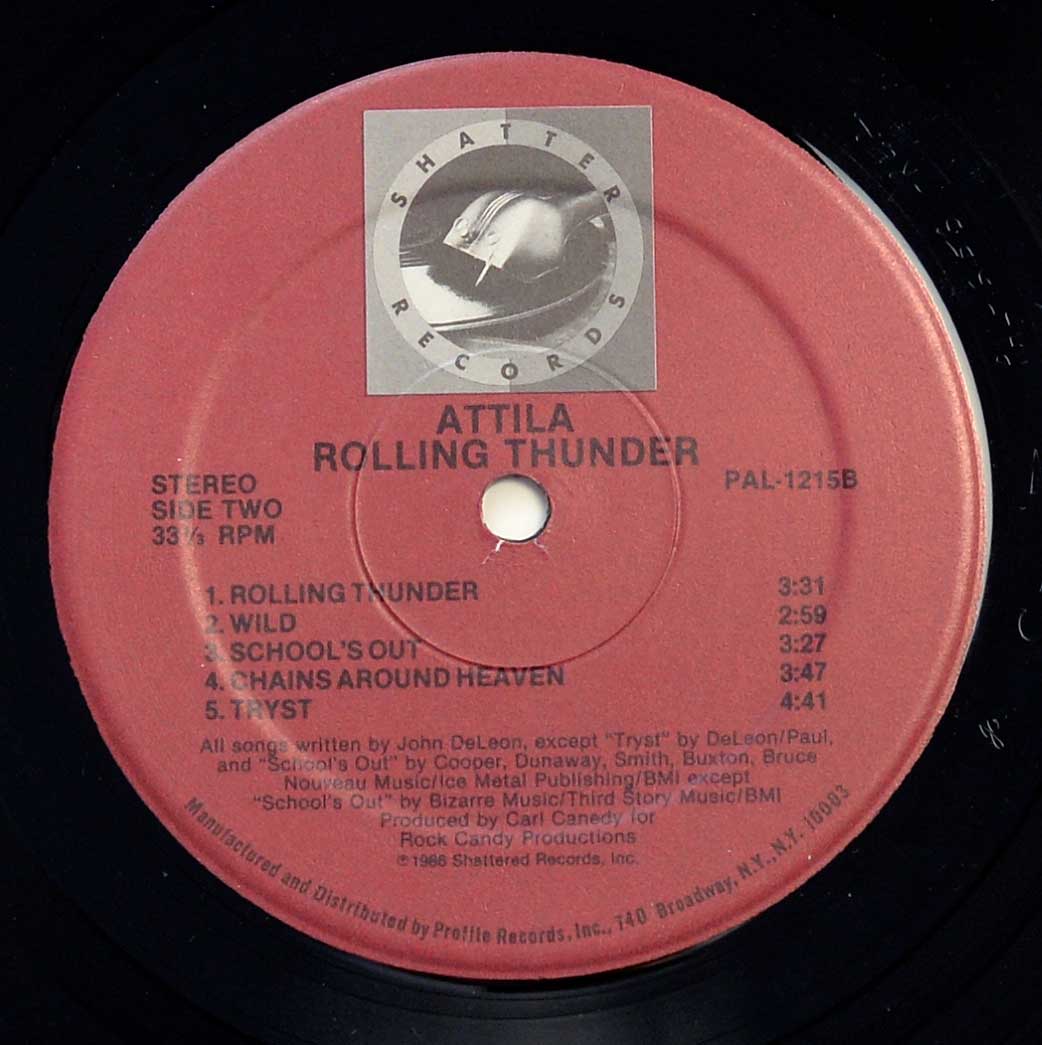 Close-up of the ATTILA - Rolling Thunder (USA) Side Two vinyl record label. The label is red with black text, featuring the 'Shatter Records' logo at the top—a metallic, shattered helmet inside a circular frame. The band's name, ATTILA, is displayed in bold uppercase letters, followed by the album title, Rolling Thunder. Below, track listings include 'Rolling Thunder,' 'Wild,' 'School’s Out,' 'Chains Around Heaven,' and 'Tryst,' with corresponding runtimes. Additional production and copyright information is printed at the bottom in smaller text.
