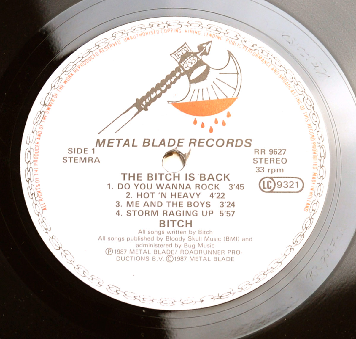 Close-up of Side One record label from 'The Bitch is Back' by Bitch (1987). The Metal Blade Records label features the iconic blood-dripping axe logo at the top. Printed in bold, the album title and tracklist include 'Do You Wanna Rock,' 'Hot 'N Heavy,' 'Me and the Boys,' and 'Storm Raging Up.' The catalog number (RR 9627) and speed (33 rpm) are displayed along with the LC 9321 logo. The outer rim carries a copyright warning, and a spindle hole is present at the center.