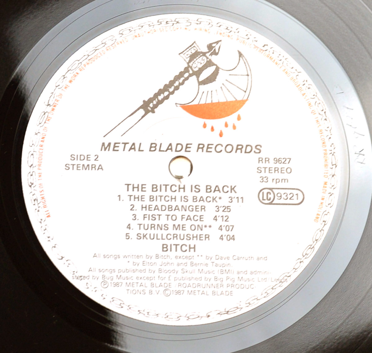 Close-up of Side Two record label from 'The Bitch is Back' by Bitch (1987). The Metal Blade Records label features the signature blood-dripping axe logo. The tracklist includes 'The Bitch is Back,' 'Headbanger,' 'Fist to Face,' 'Turns Me On,' and 'Skullcrusher.' The catalog number (RR 9627), stereo format, and LC 9321 code are displayed. Fine-print copyright information and a central spindle hole complete the label design.