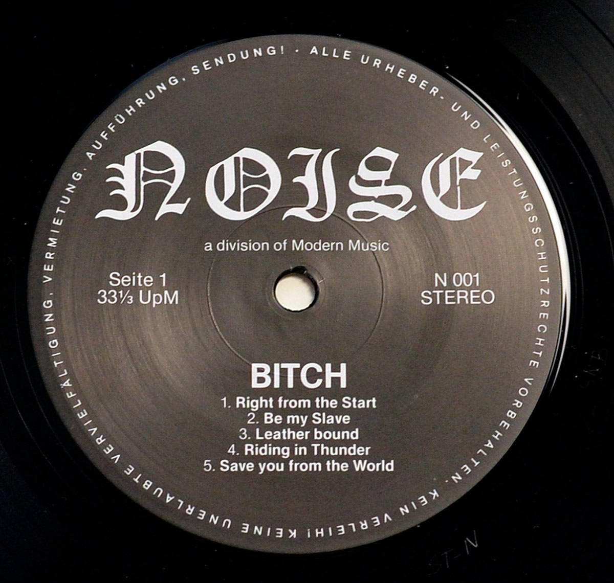 Close-up of Side One label of BITCH - Be My Slave vinyl LP. The black label features the NOISE Records logo in bold gothic font, along with track listing for Side One: 'Right From The Start,' 'Be My Slave,' 'Leatherbound,' 'Riding in Thunder,' and 'Save You From The World.' The catalog number N 001 and stereo format are displayed.