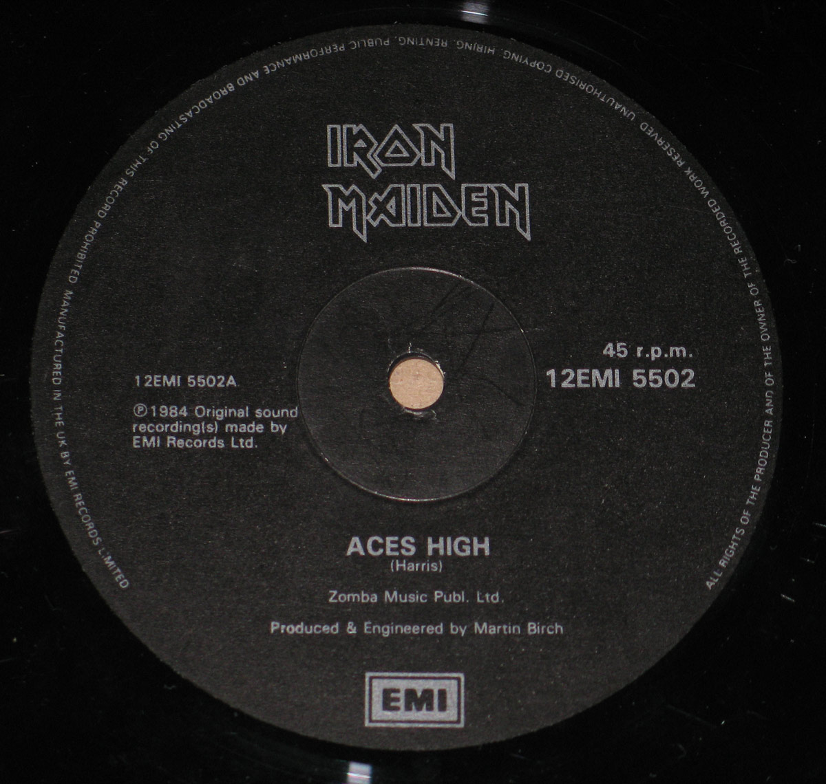 Close up of Side One label for Iron Maiden Aces High UK maxi 12-inch vinyl showing a matte black EMI label with silver-grey text. The stylized Iron Maiden band logo sits at the top, catalogue numbers 12EMI 5502 and 12EMI 5502A appear left and right of the spindle hole, and the song title Aces High is printed in the lower half above the EMI box logo. Fine circular rim text runs around the outer edge, and the glossy black vinyl grooves surround the label.