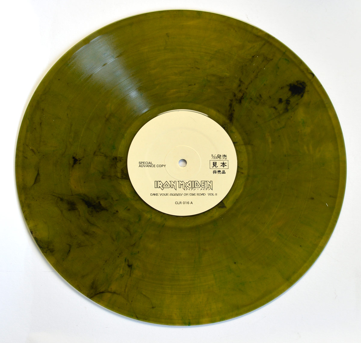 A translucent olive-green marbled vinyl record of Iron Maiden – Take Your Mummy on the Road Vol. II. The record has a smoky, dark marbling effect, giving it a unique and eerie appearance. The center label is off-white with black text, featuring the Iron Maiden logo along with the Japanese text アイアン・メイデン (Iron Maiden). Below, the album title is printed, along with the catalog number CLR 016 A, indicating Side A. The label also states Special Advance Copy and includes Japanese characters 見本 (mihon), meaning “sample” or “not for sale,” suggesting it is a promotional or test pressing. The design complements the album’s samurai-horror aesthetic.