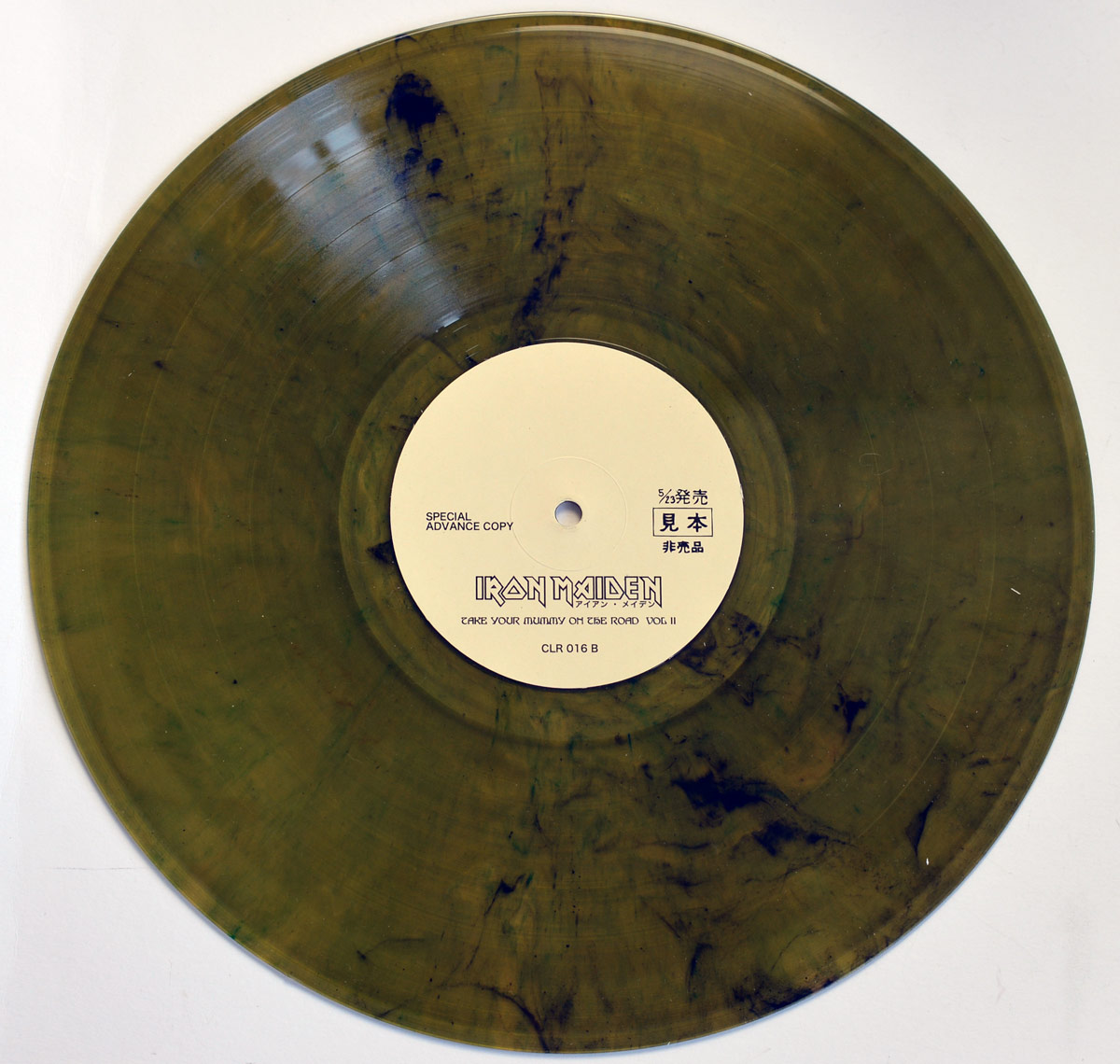 A translucent olive-green marbled vinyl record of Iron Maiden – Take Your Mummy on the Road Vol. II, Side B. The record has a smoky, dark marbling effect, creating a unique and eerie appearance. The center label is off-white with black text, featuring the Iron Maiden logo along with the Japanese text アイアン・メイデン (Iron Maiden). Below, the album title is printed, followed by the catalog number CLR 016 B, designating this as Side B. The label also states Special Advance Copy and includes Japanese characters 見本 (mihon), meaning “sample” or “not for sale,” indicating a promotional or test pressing. The design complements the album’s samurai-horror aesthetic