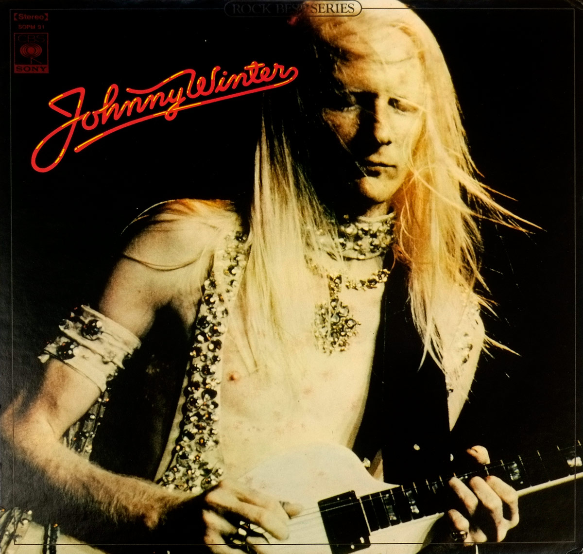 Close-up of Johnny Winter on the front cover of the Japan-only LP 'The Best of Johnny Winter'. He plays a white Gibson Firebird with his long platinum hair flowing. His bare chest is adorned with ornate jewelry under a spotlight.
