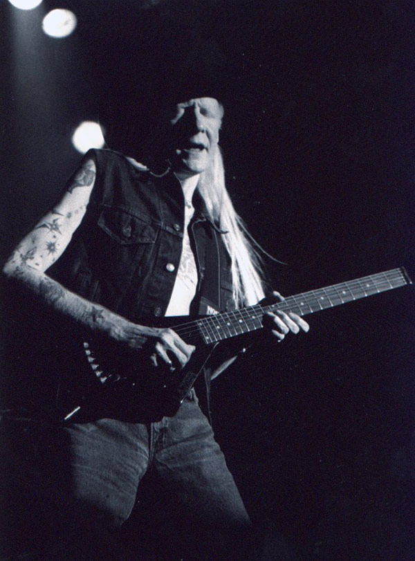Black and white photograph of Johnny Winter mid-performance. He wears a black cowboy hat, a sleeveless denim vest, and jeans. His arms are tattooed, and his intense facial expression shows deep focus. The stage lights shine behind him, casting dramatic shadows as he plays a uniquely shaped Hondo electric guitar with a sharp, angular body. https://vinyl-records.nl/