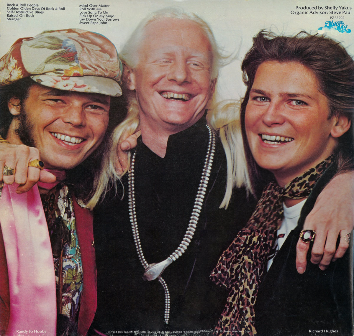 Back cover photo of Johnny Winter smiling broadly between two bandmates, Randy Jo Hobbs on the left and Richard Hughes on the right. All three stand close together with arms around each other’s shoulders. Winter wears a dark jacket and a long silver necklace. Hobbs wears a patterned cap, layered necklaces and a pink scarf. Hughes wears a dark jacket with a leopard-print scarf. Track listings and production credits appear at the top, with the Blue Sky Records logo in the upper right.
