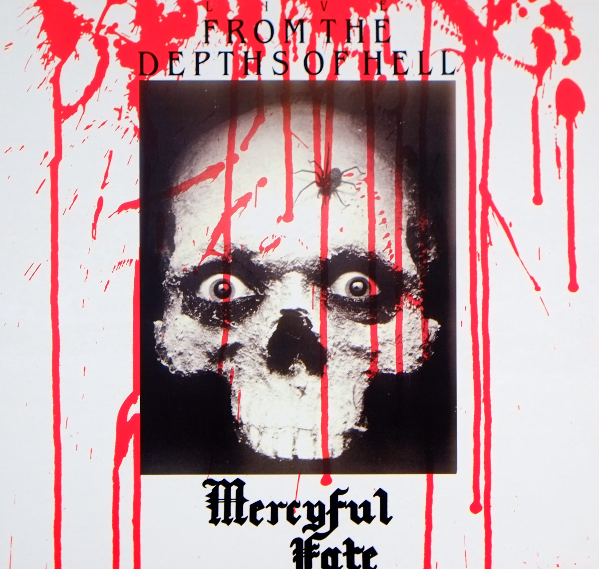 Album cover of “From the Depths of Hell” by Mercyful Fate. It features a black-and-white image of a skull-painted face with wide, eerie eyes and a spider on the forehead. Red paint, resembling dripping blood, covers the white background. The album title appears at the top in gothic font, while the band’s name, Mercyful Fate, is displayed at the bottom in blackletter typography