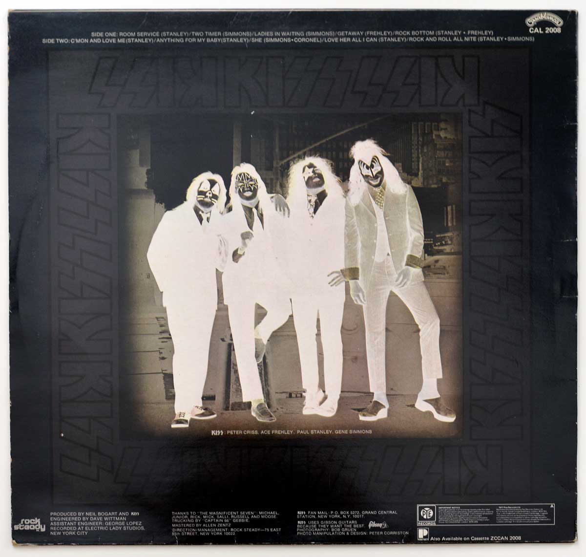 Back cover of KISS’s 1975 album 'Dressed to Kill,' showing the continuation of the same New York City street scene from the front cover, now reversed in perspective. The four band members — Gene Simmons, Paul Stanley, Ace Frehley, and Peter Criss — still stand in their formal business suits and trademark makeup, their poses slightly altered, suggesting a moment between shots. The muted tones and simple background highlight the raw, unvarnished aesthetic of 1970s rock imagery. Around the border, the repeating KISS logo remains embossed in gray-black, while the Casablanca Records logo appears faintly in the upper left corner, marking the label that produced the album. The composition reflects the label’s tight production budget and no-frills presentation, yet captures the band’s confident attitude and ironic humor.