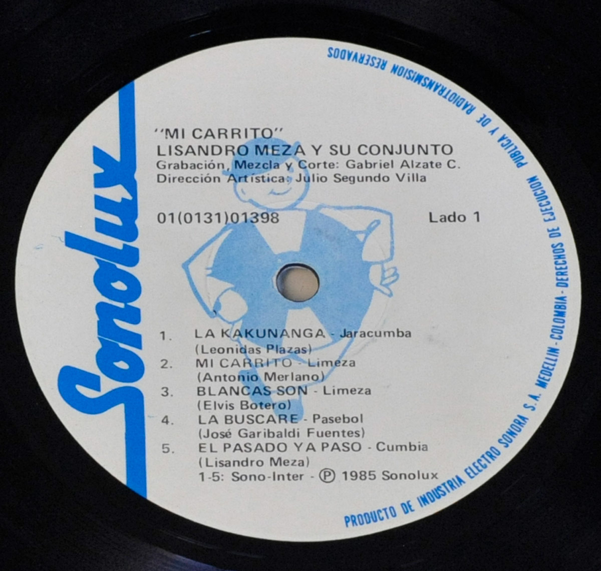 Close-up of Side 1 label of 'Mi Carrito' by Lisandro Meza. The label is white with blue text and features the Sonolux logo in bold blue on the left. A light blue illustration of a cartoon character playing an instrument is in the background. The album title and artist name are at the top, followed by production credits for Gabriel Alzate C. and artistic direction by Julio Segundo Villa. The catalog number 01(0131)01398 is printed below, along with the tracklist for Side 1, which includes five songs with their respective composers. The outer rim has small blue text indicating copyright information and rights management. The vinyl’s black edge frames the label.