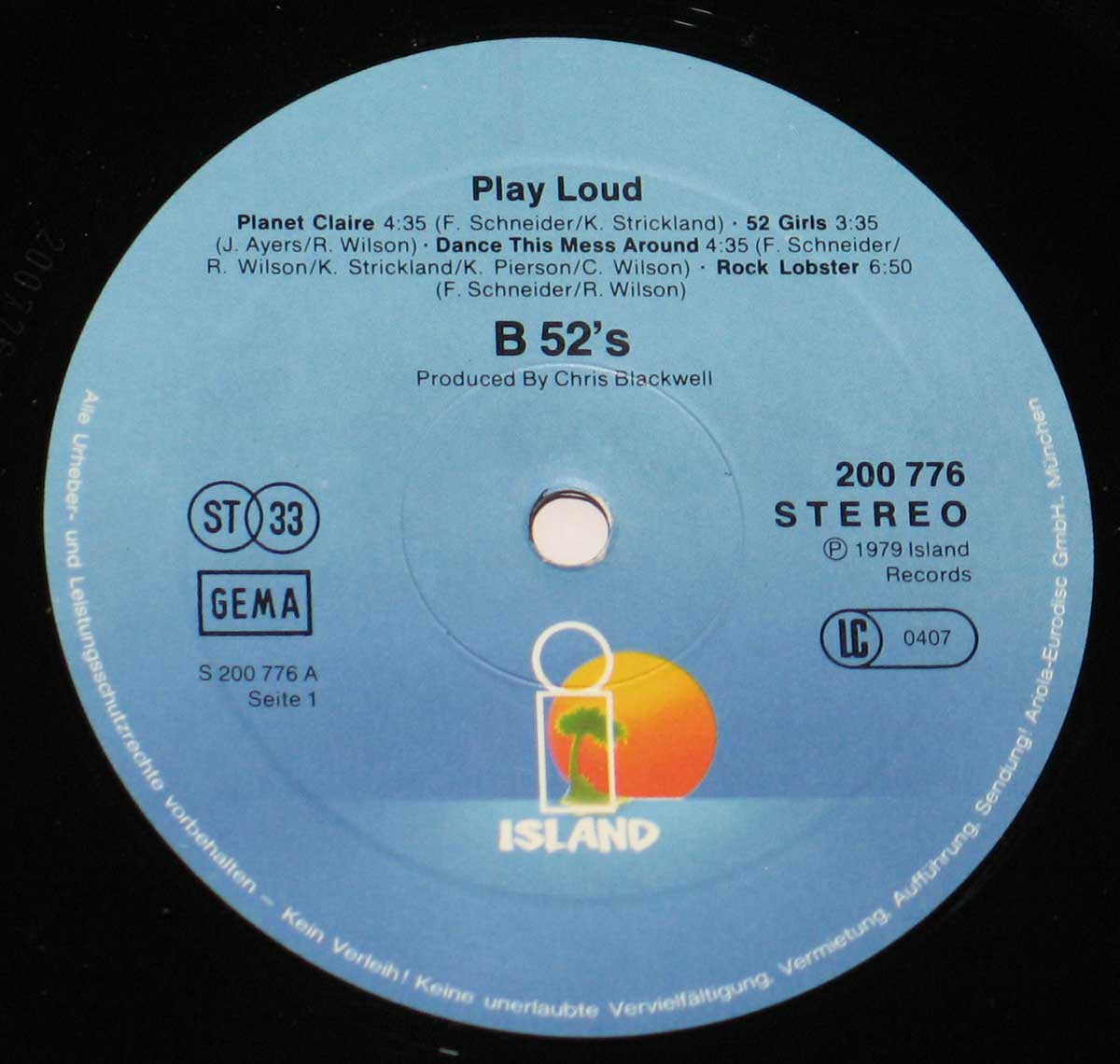 Close-up of Side One record label for The B-52’s self-titled debut album (German release). The circular blue label carries the classic Island Records palm tree logo at the bottom — a yellow and orange sunset over a small green palm — with the word 'ISLAND' printed in white capitals beneath it. The text above reads 'Play Loud' followed by the four tracks on Side One: 'Planet Claire,' '52 Girls,' 'Dance This Mess Around,' and 'Rock Lobster,' each with songwriting credits. The upper text also lists 'Produced by Chris Blackwell.' On the right, the catalog number '200 776' and 'STEREO' marking appear above the copyright line '(P) 1979 Island Records' and the LC code '0407.' On the left are the GEMA logo and the ST33 speed symbol, all printed in crisp black type. Around the edge runs fine white German copyright text, completing a clean late-1970s design.