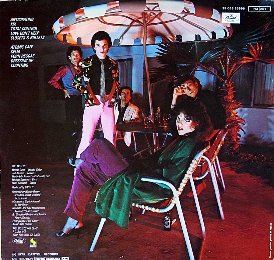 Back cover of The Motels’ 1979 self-titled album featuring the band gathered at a motel courtyard patio table under a striped umbrella, with dramatic lighting in blue, pink, and red hues highlighting Martha Davis seated prominently in the foreground.
