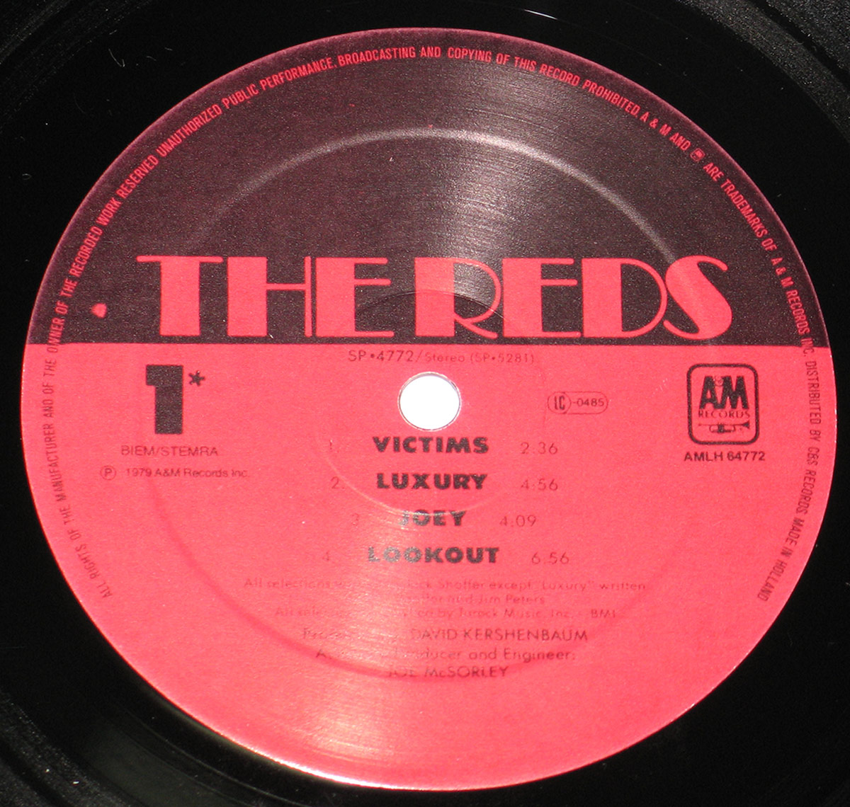 Close-up of The Reds' 1979 vinyl record label from A&M Records. The label is red with black text, featuring the band's name in bold, stylized font at the top. It includes track listings such as 'Victims,' 'Luxury,' 'Joey,' and 'Lookout,' along with production credits for David Kershenbaum. The record is black, with the grooves reflecting light.