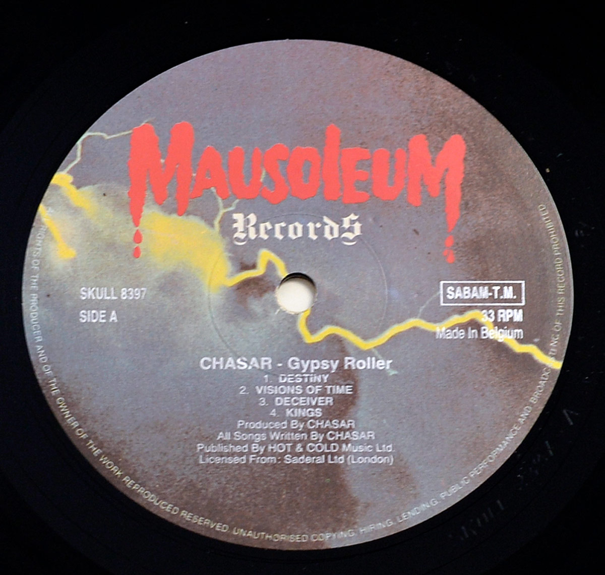 Close-up photo of Side A label of CHASAR's 1987 album 'Gypsy Roller,' released under Mausoleum Records. The label design features a dark, stormy sky with yellow lightning bolts and the bold red 'Mausoleum Records' logo dripping like blood at the top. The tracklist for Side A includes 'Destiny,' 'Visions of Time,' 'Deceiver,' and 'Kings.' Additional credits indicate production by CHASAR, with licensing from Saderal Ltd (London) and publishing through HOT & COLD Music Ltd. The vinyl is pressed in Belgium at 33 RPM.