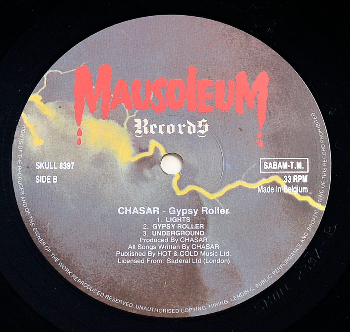 Close-up photo of Side B label of CHASAR's 1987 album 'Gypsy Roller,' released under Mausoleum Records. The label features a stormy sky background with striking yellow lightning bolts, while the bold red 'Mausoleum Records' logo drips ominously at the top. The tracklist for Side B includes 'Lights,' 'Gypsy Roller,' and 'Underground.' Additional credits list production by CHASAR, with publishing through HOT & COLD Music Ltd and licensing from Saderal Ltd (London). The vinyl is pressed in Belgium at 33 RPM.