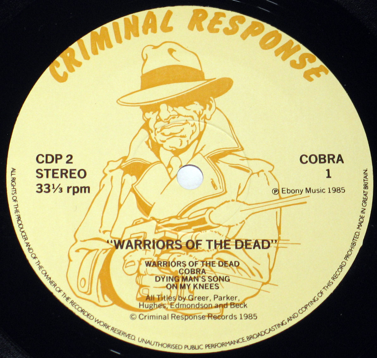 Close-up of Side One label of Cobra's 'Warriors of the Dead' vinyl LP. The label is beige with a bold 'Criminal Response' logo at the top. It features an illustration of a trench-coated detective with a fedora, holding a sawed-off shotgun. Tracklist includes 'Warriors of the Dead,' 'Cobra,' 'Dying Man’s Song,' and 'On My Knees.' Criminal Response Records, Ebony Music, 1985.