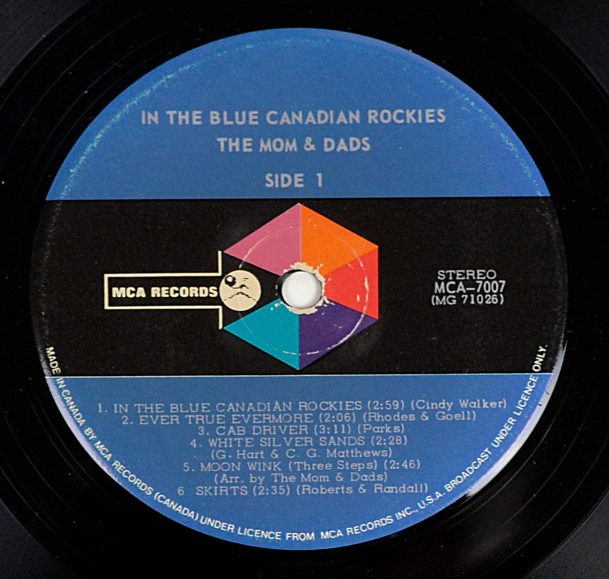 Side 1 of the 12-inch vinyl LP THE MOM AND DADS - In The Blue Canadian Rockies. The MCA Records label features a colorful geometric prism design on black with blue and purple upper half. White text lists the album title, band name, and six track titles including In The Blue Canadian Rockies, Ever True Evermore, and Cab Driver.