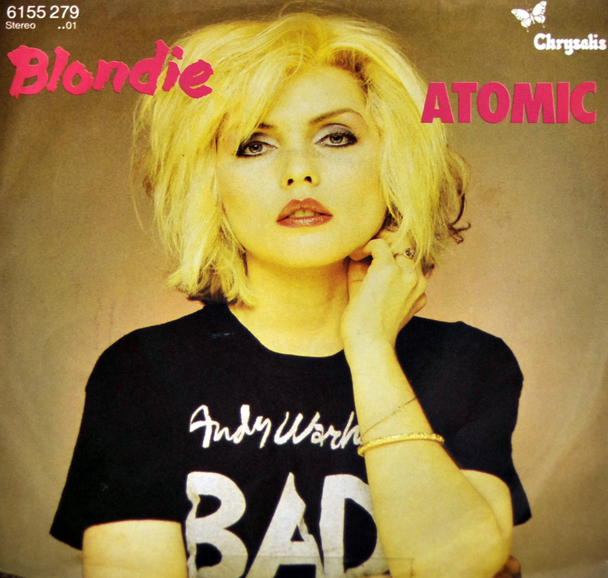Cover art for Blondie's single Atomic featuring Debbie Harry with platinum blonde hair wearing a black T-shirt that reads Andy Warhol's BAD She poses confidently against a muted background with Blondie in pink text on the left Atomic in bold pink on the right and the Chrysalis Records butterfly logo in the top right corner https://vinyl-records.nl