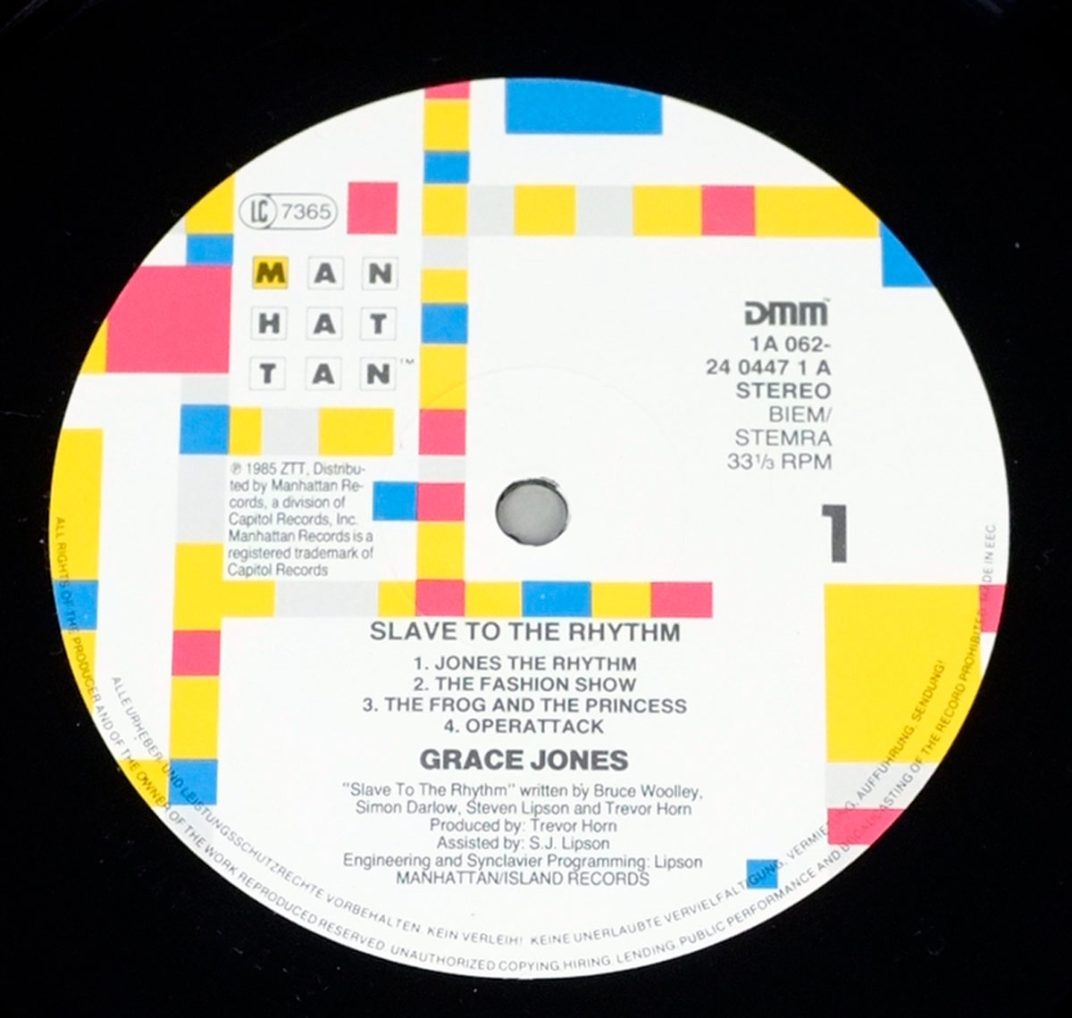 Close-up of the Side One record label for Grace Jones - Slave to the Rhythm. The circular label is white with bold geometric design elements in red, yellow, blue, and pink squares and rectangles arranged around the edges. The Manhattan Records logo appears at the top left, with the letters MAN HAT TAN stacked in black squares. To the right, details include catalog number 1A 062-24 0447 1 A, Stereo, BIEM/STEMRA, DMM, and playback speed 33 1/3 RPM. Below the center hole, the album title Slave to the Rhythm is printed in bold capitals, followed by the four Side One tracks: Jones the Rhythm, The Fashion Show, The Frog and the Princess, and Operattack. The artist’s name Grace Jones is centered below in bold. Writing and production credits name Bruce Woolley, Simon Darlow, Steven Lipson, and Trevor Horn, with Horn listed as producer and S.J. Lipson as assistant. Additional credits for engineering and Synclavier programming by Lipson are included. The bottom edge states Manhattan/Island Records along with copyright and distribution details.