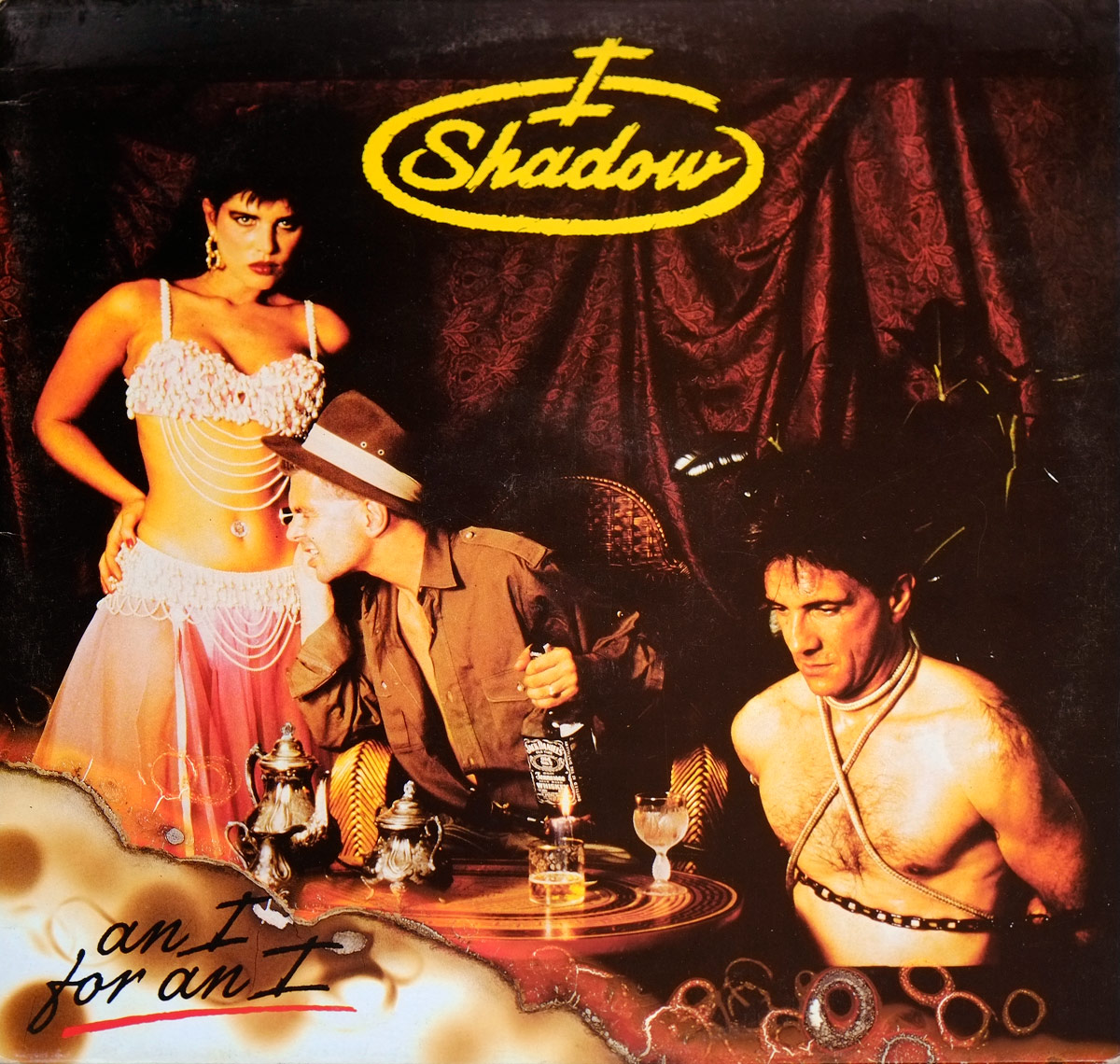 Front cover of I SHADOW – An I For An I showing a theatrical and provocative tableau with a belly dancer, a seated man holding a bottle of Jack Daniel's, and a shirtless man tied with rope in an exotic, dimly lit setting. https://vinyl-records.nl/